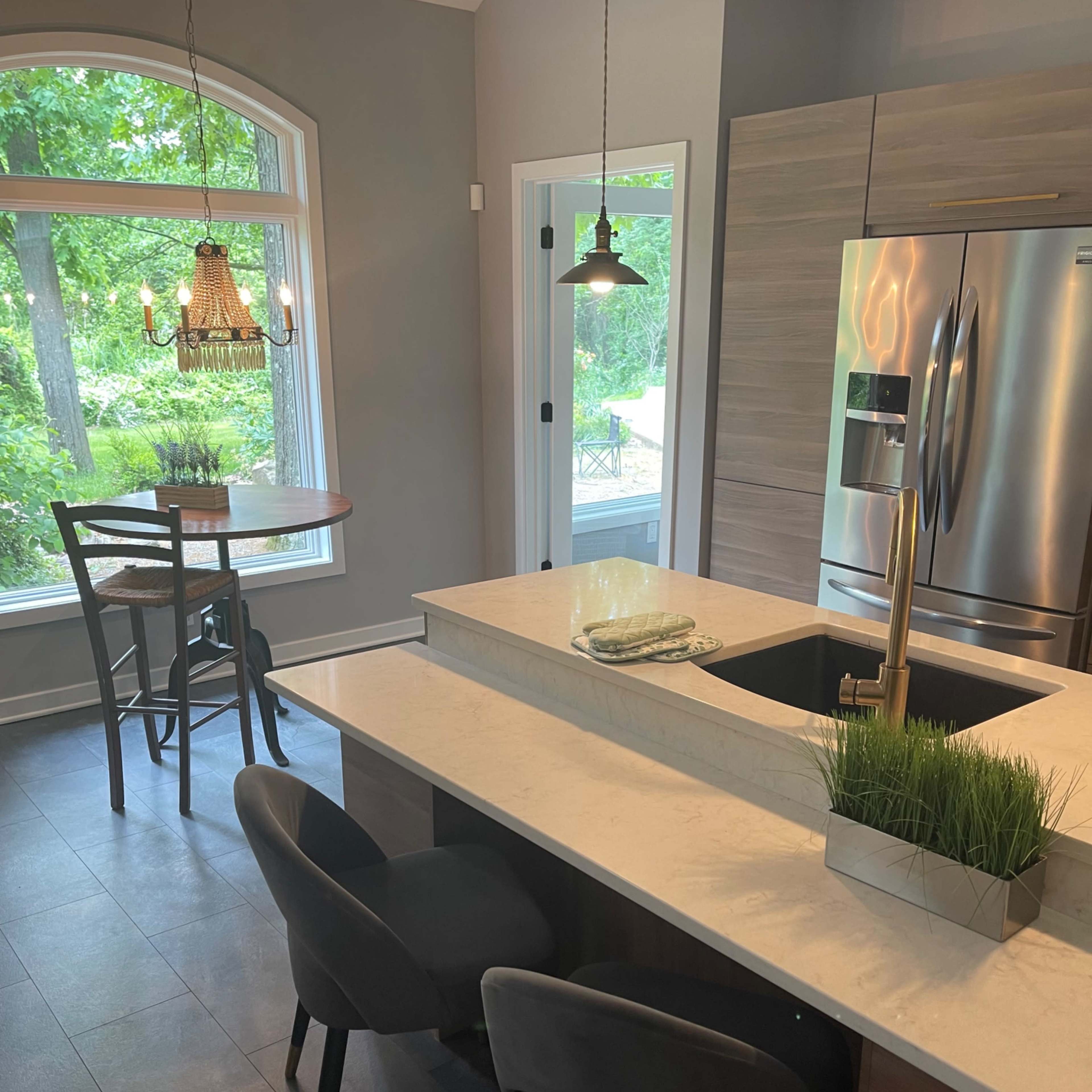 A modern kitchen with a marble island, two gray chairs, a dining table for two with pendant lighting, and large windows overlooking a green outdoor area.