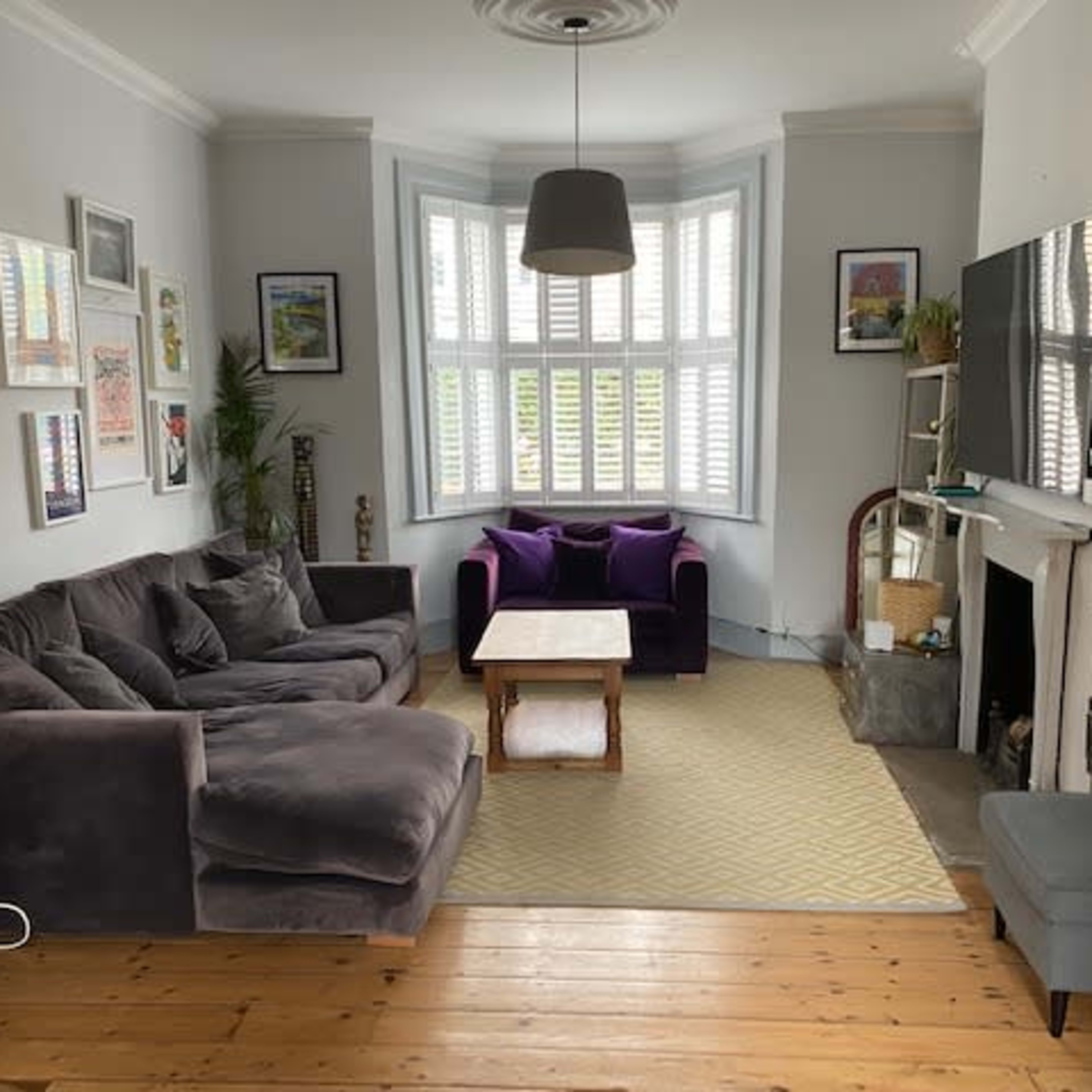 The living room features a gray sectional sofa, two purple armchairs, a hardwood floor, and a large bay window with white shutters.