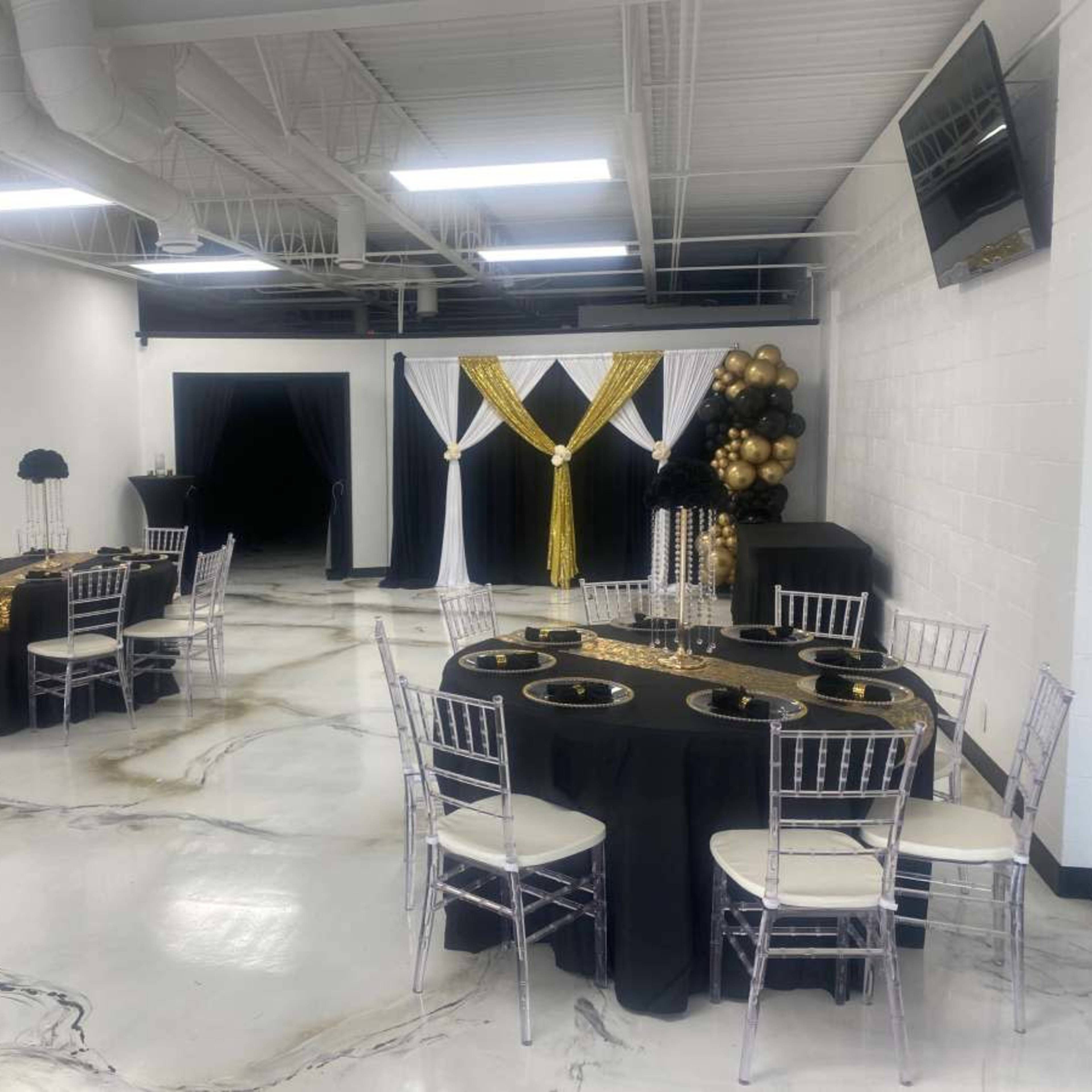 A decorated event space with several tables set with black and gold tablecloths, elegant chairs, and a backdrop featuring gold and black decor.