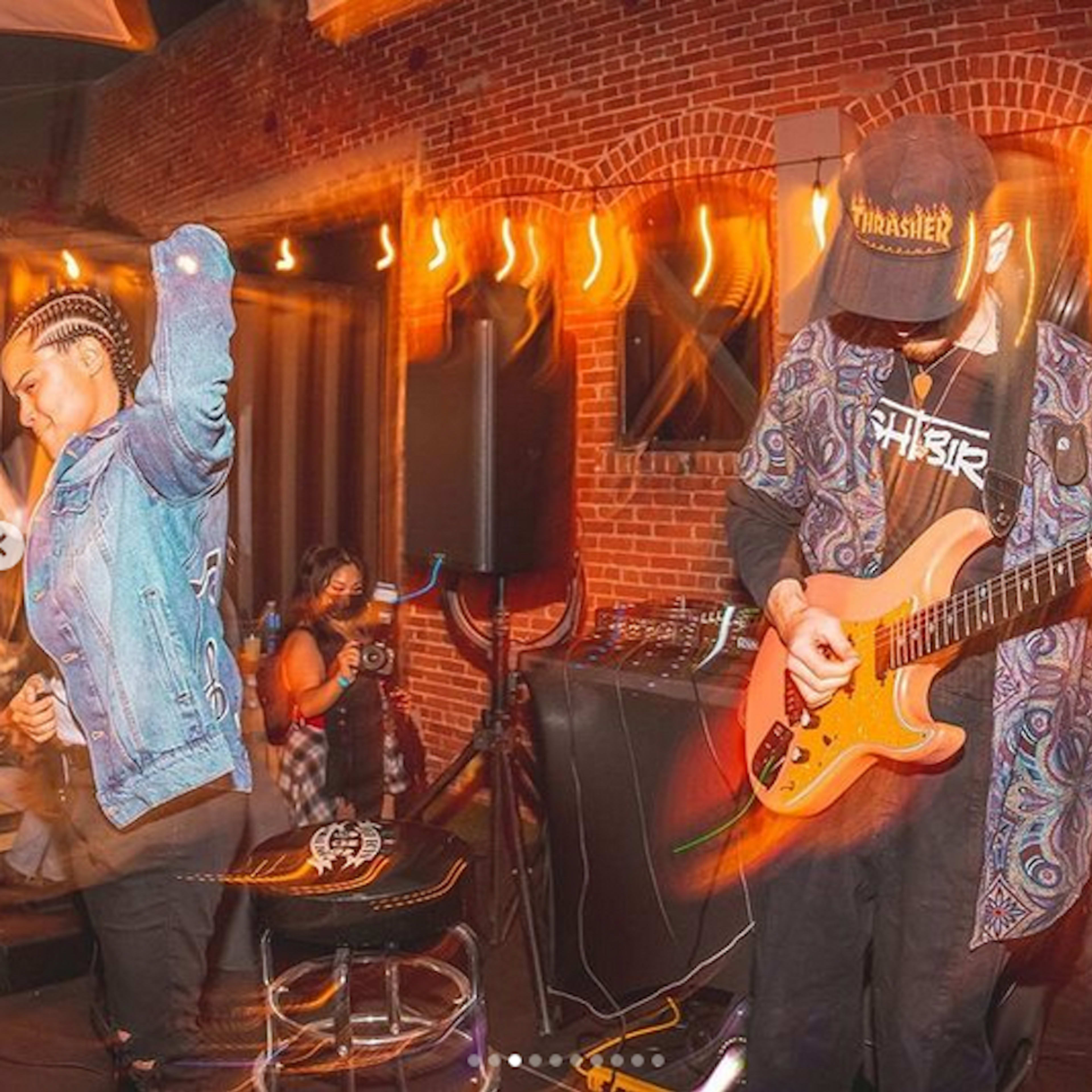 A musician plays an orange guitar on stage while a woman dances nearby in a dimly lit venue.