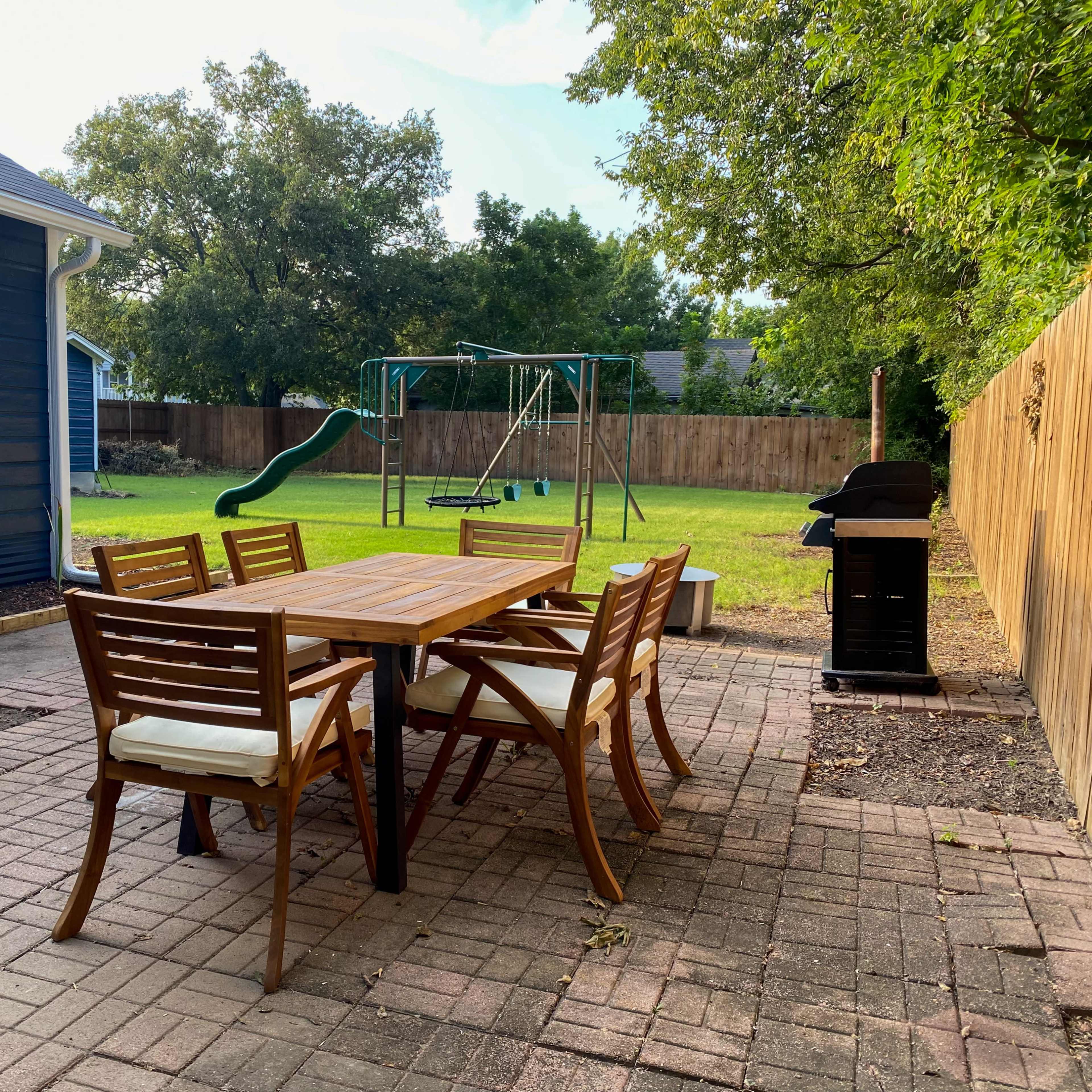 A wooden dining table with chairs is set on a brick patio, next to a swing set and a barbecue grill in a yard with a wooden fence.