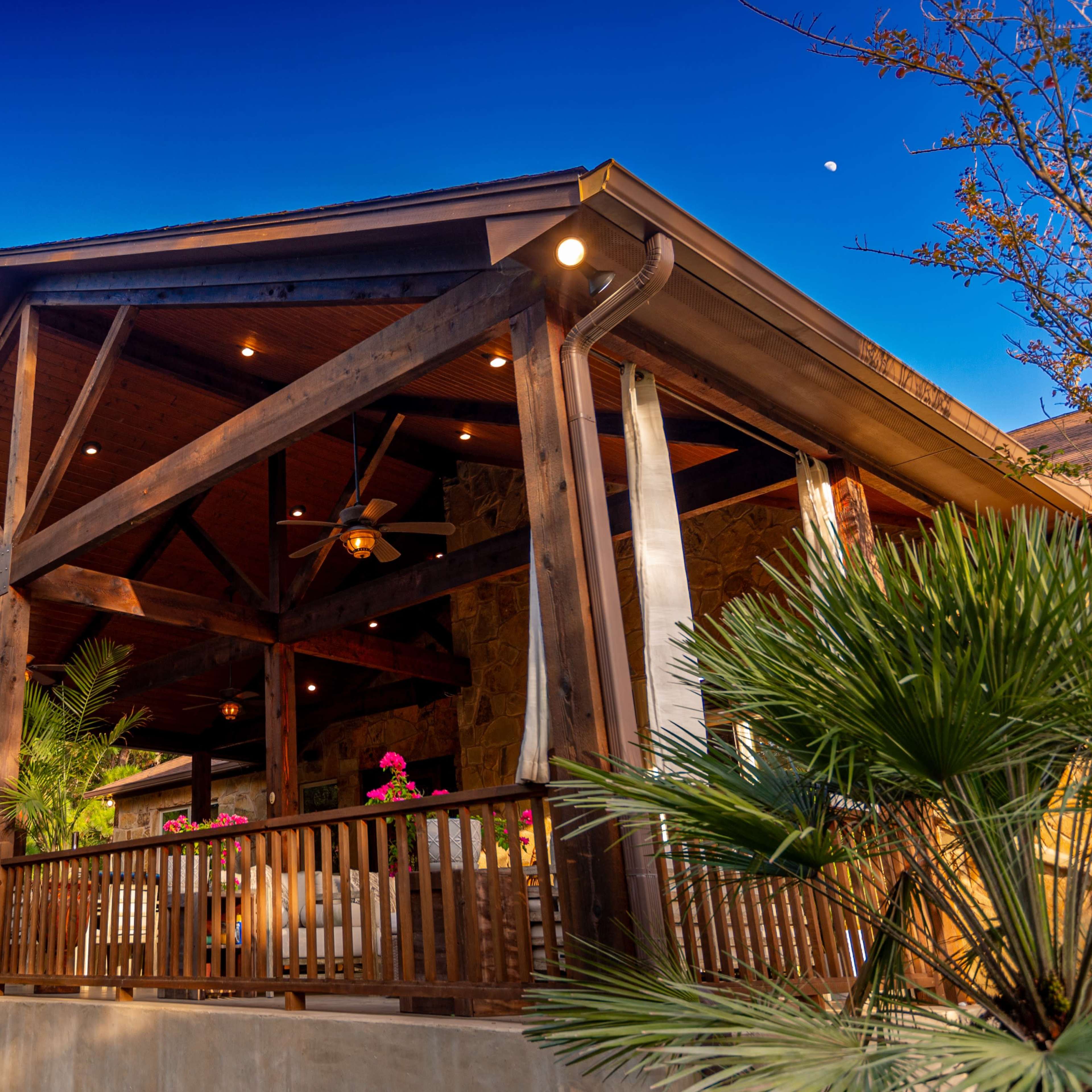 A wooden porch with a gabled roof supported by beams, surrounded by greenery and featuring potted plants.