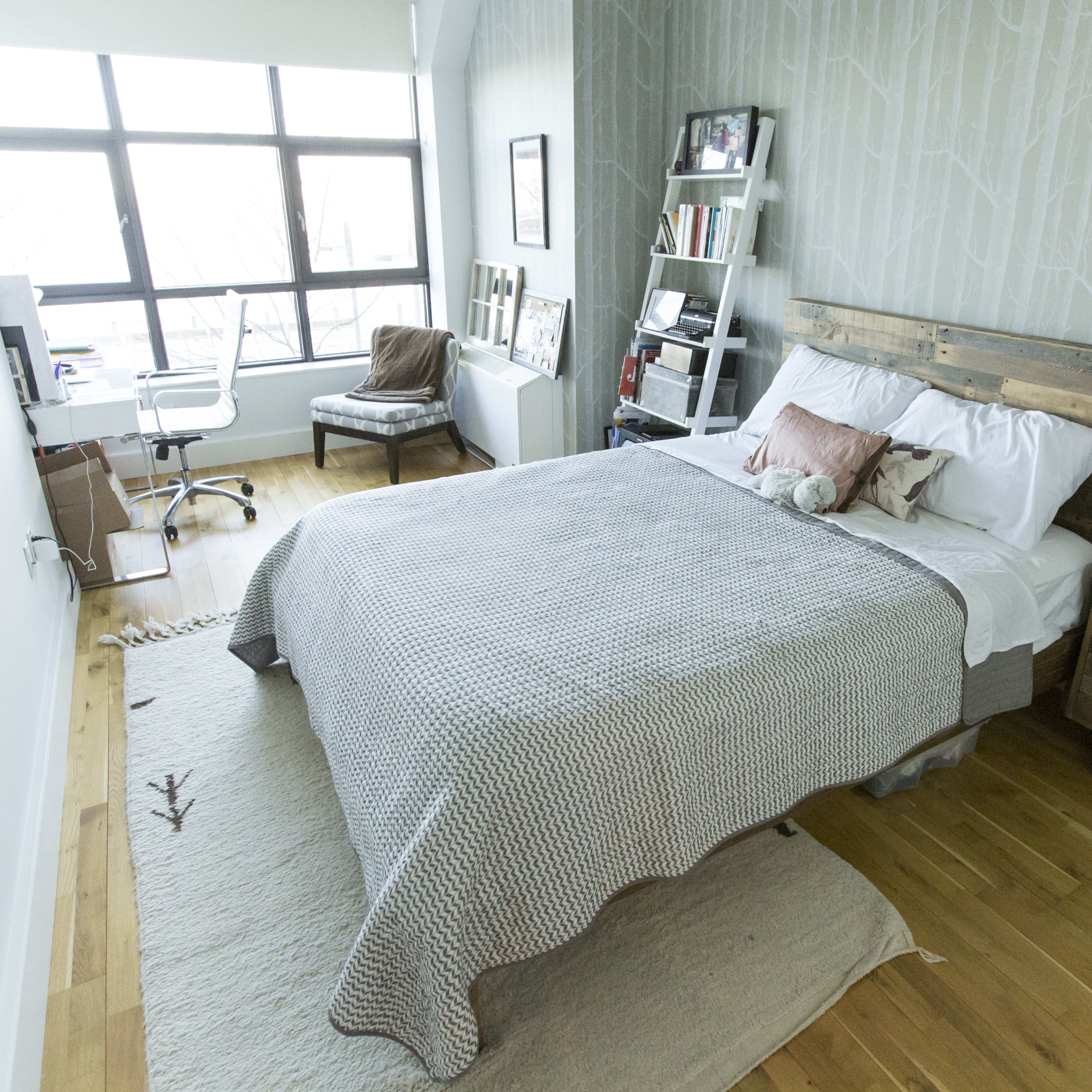 A neatly organized bedroom features a wooden bed with a light-colored blanket, a chair, a small desk, and large windows allowing natural light to enter.