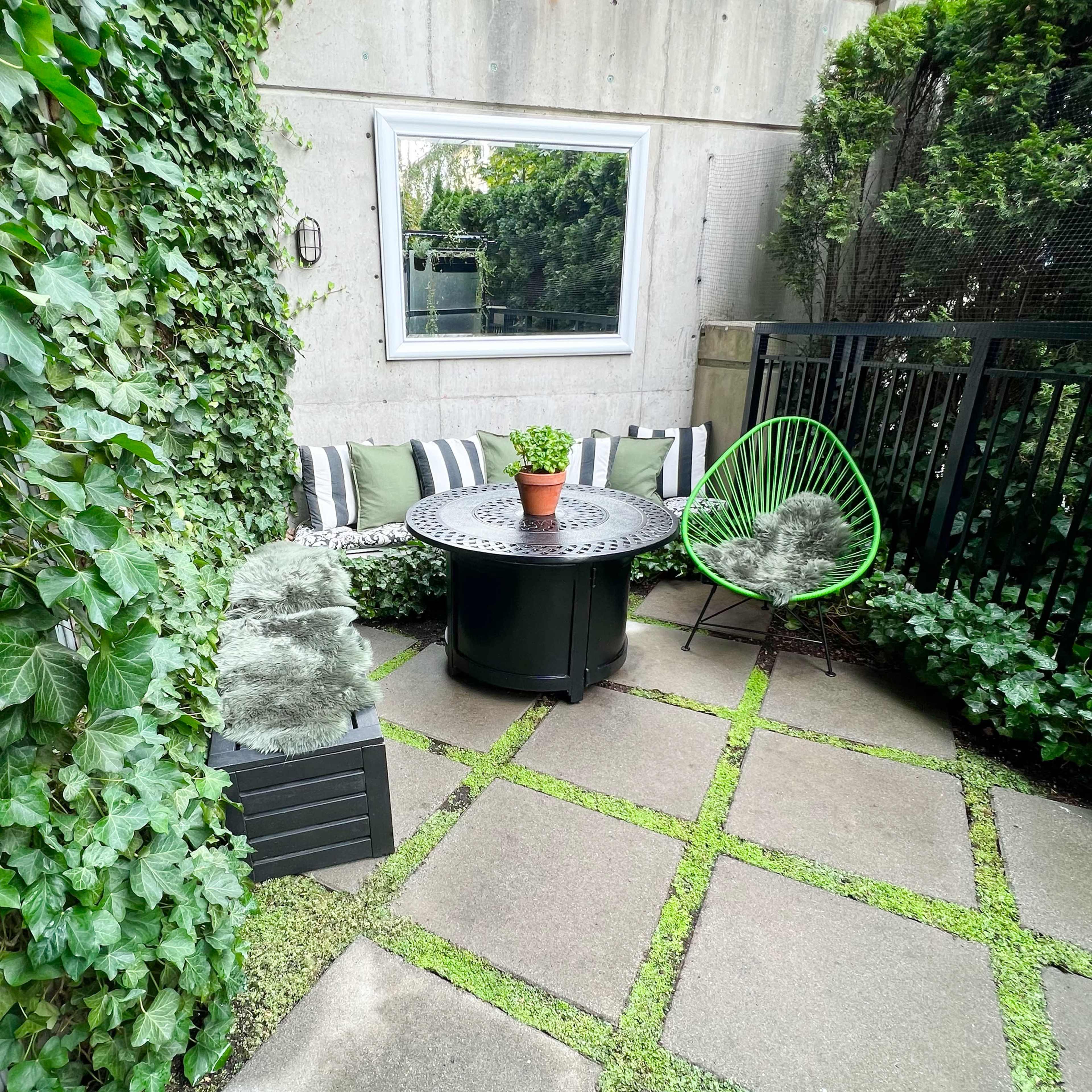 A small outdoor patio surrounded by greenery, featuring a circular table with a potted plant and two chairs.