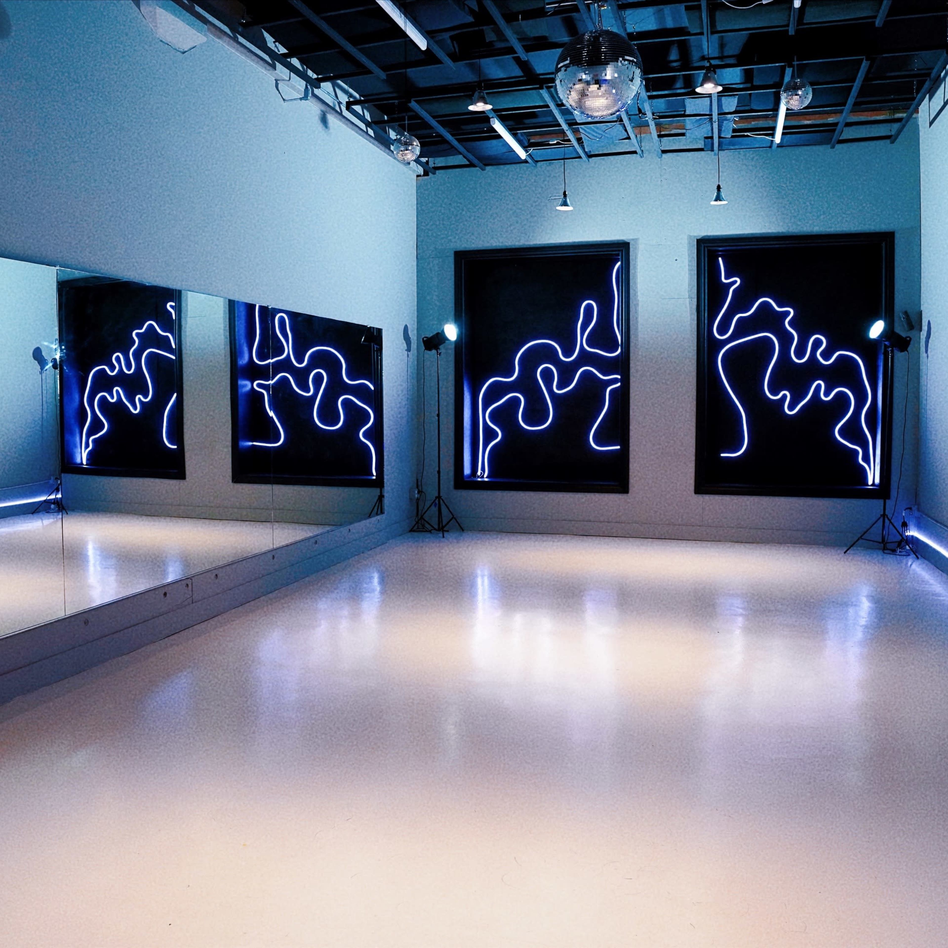 A dance studio featuring large mirrors on one wall, abstract illuminated art on the opposite wall, and a smooth white floor.