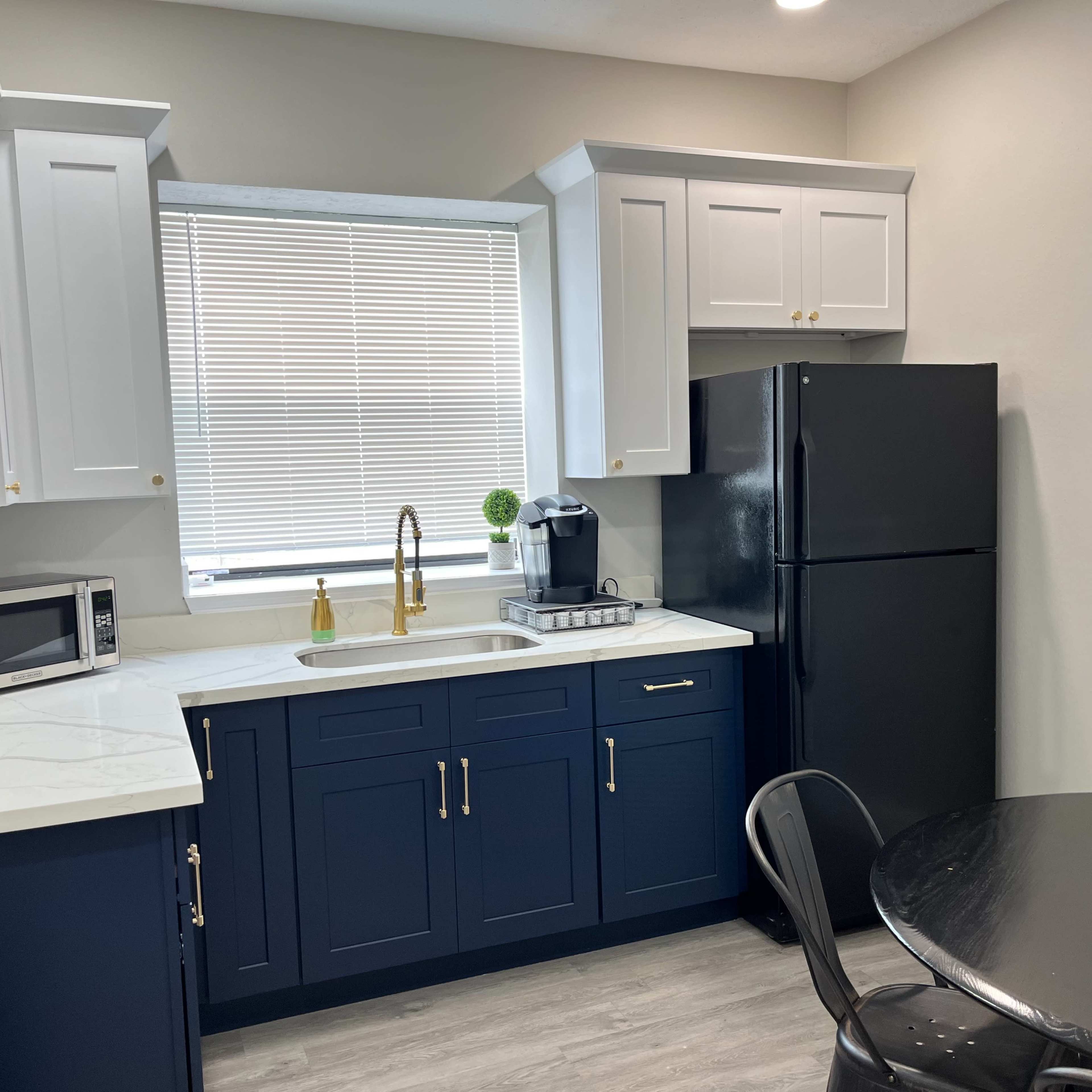 A clean kitchen featuring navy blue cabinets, a white countertop, a stainless steel sink, a black refrigerator, and a round black table.