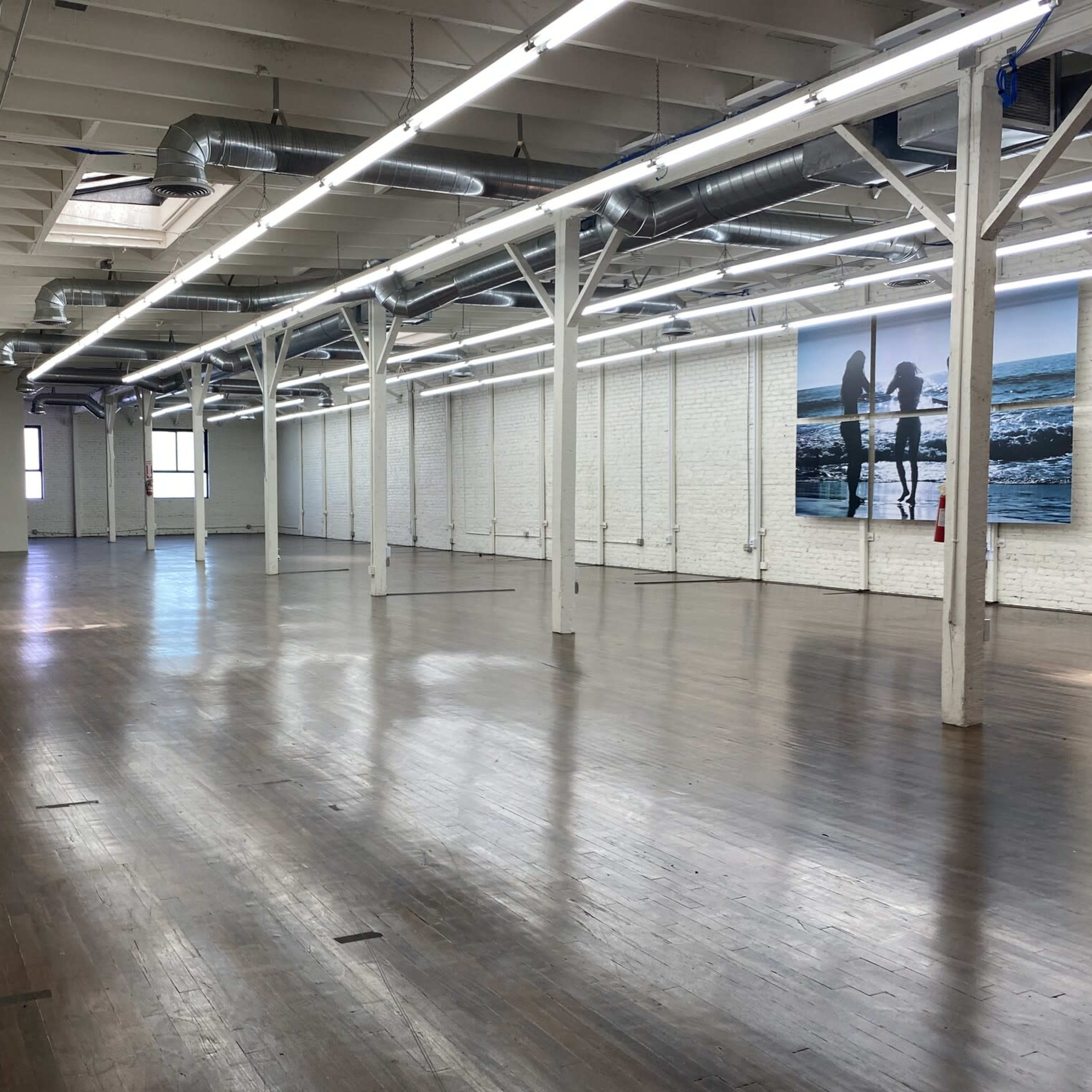 A spacious, empty loft with wooden floors and exposed beams, featuring white brick walls and large windows.