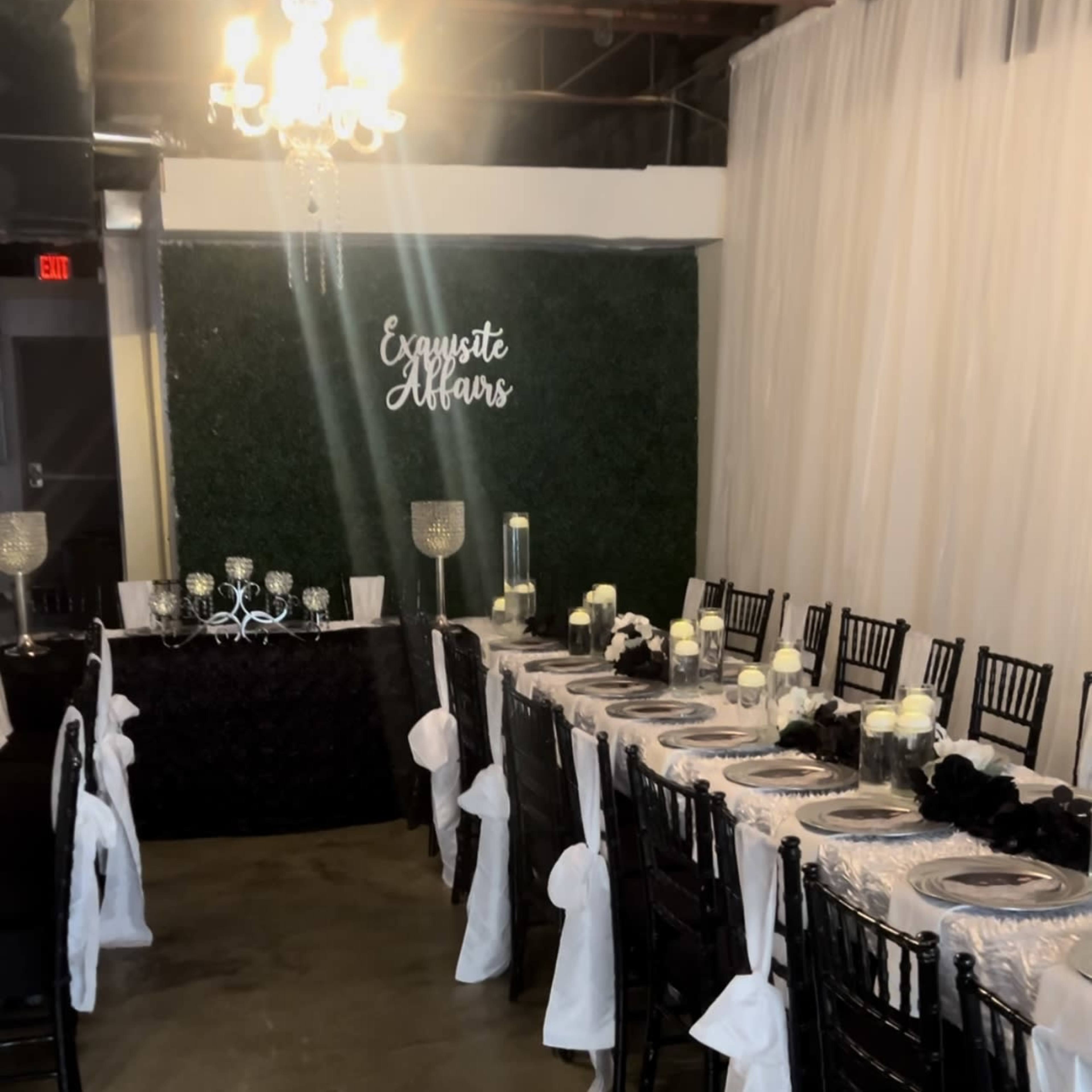 A long dining table adorned with white tablecloths and black accents is set up in a banquet space featuring greenery on the wall and a chandelier overhead.