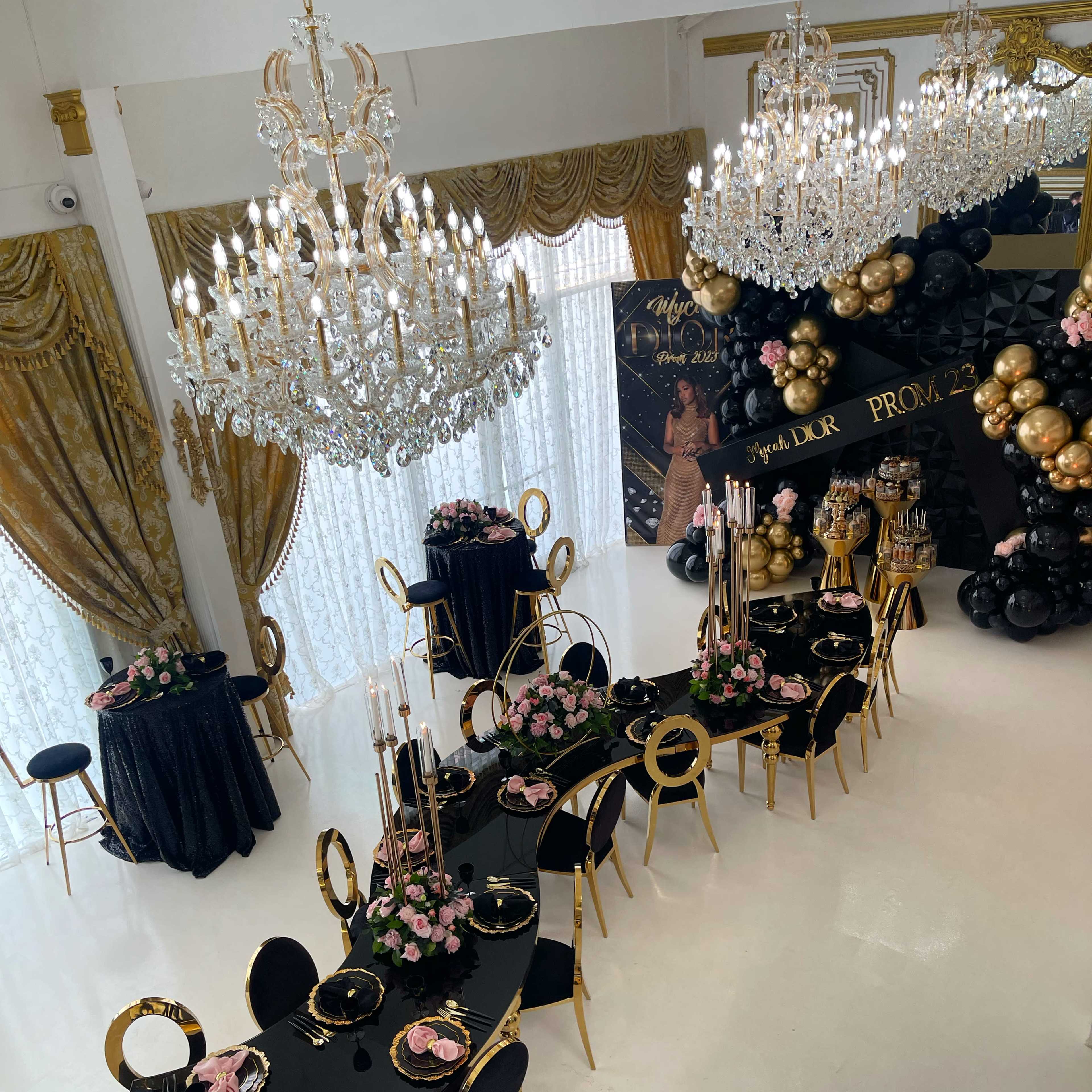 A lavish banquet hall features a long, elegantly set table with black and gold decor, surrounded by chandeliers and floral arrangements.