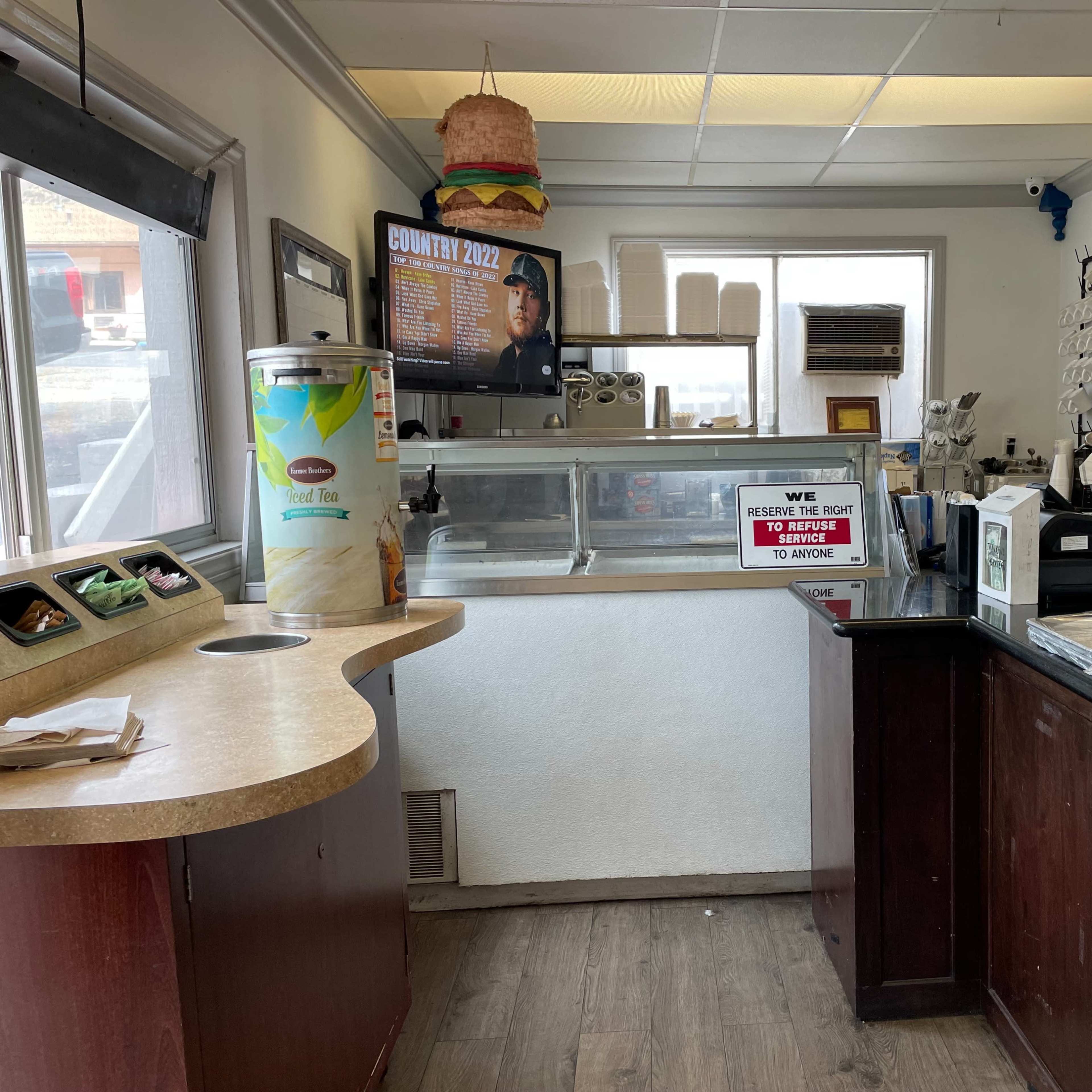 A small restaurant interior features a counter with menu displays, a cash register, and various food items visible on the shelves.