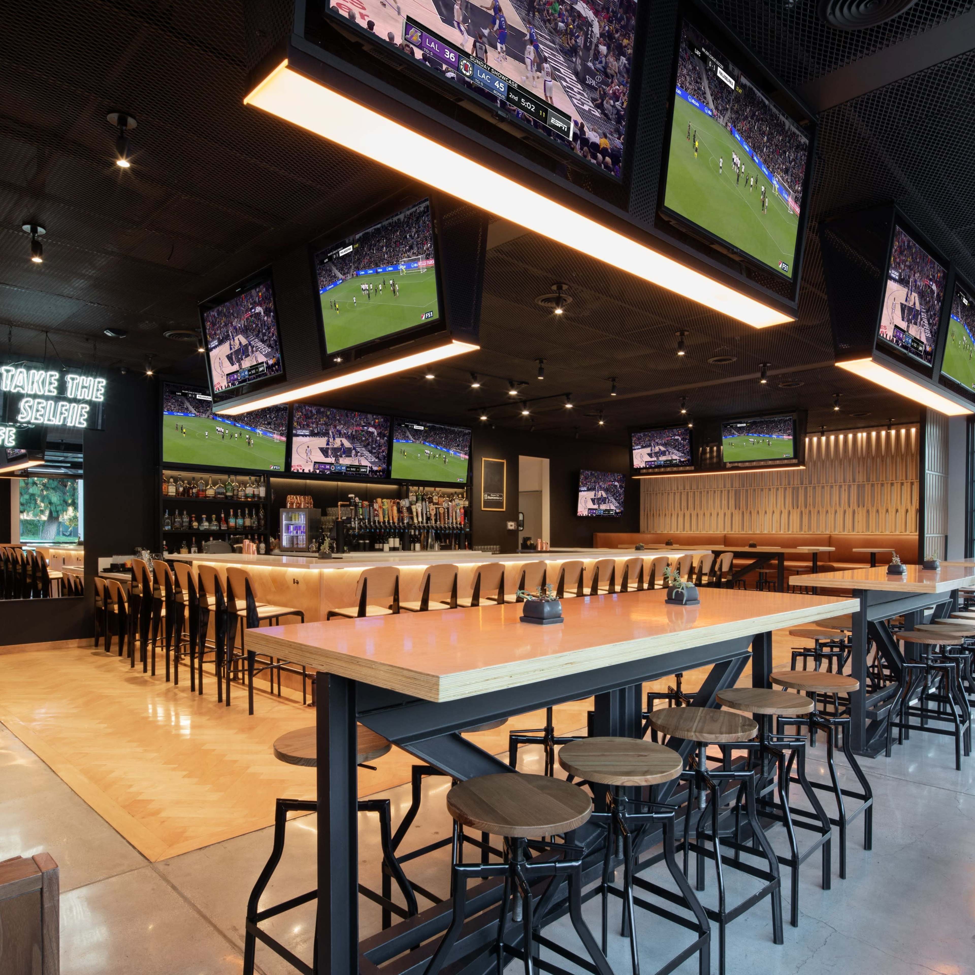 A modern sports bar with multiple screens displaying a game, featuring a long bar counter with seating and a sleek design.