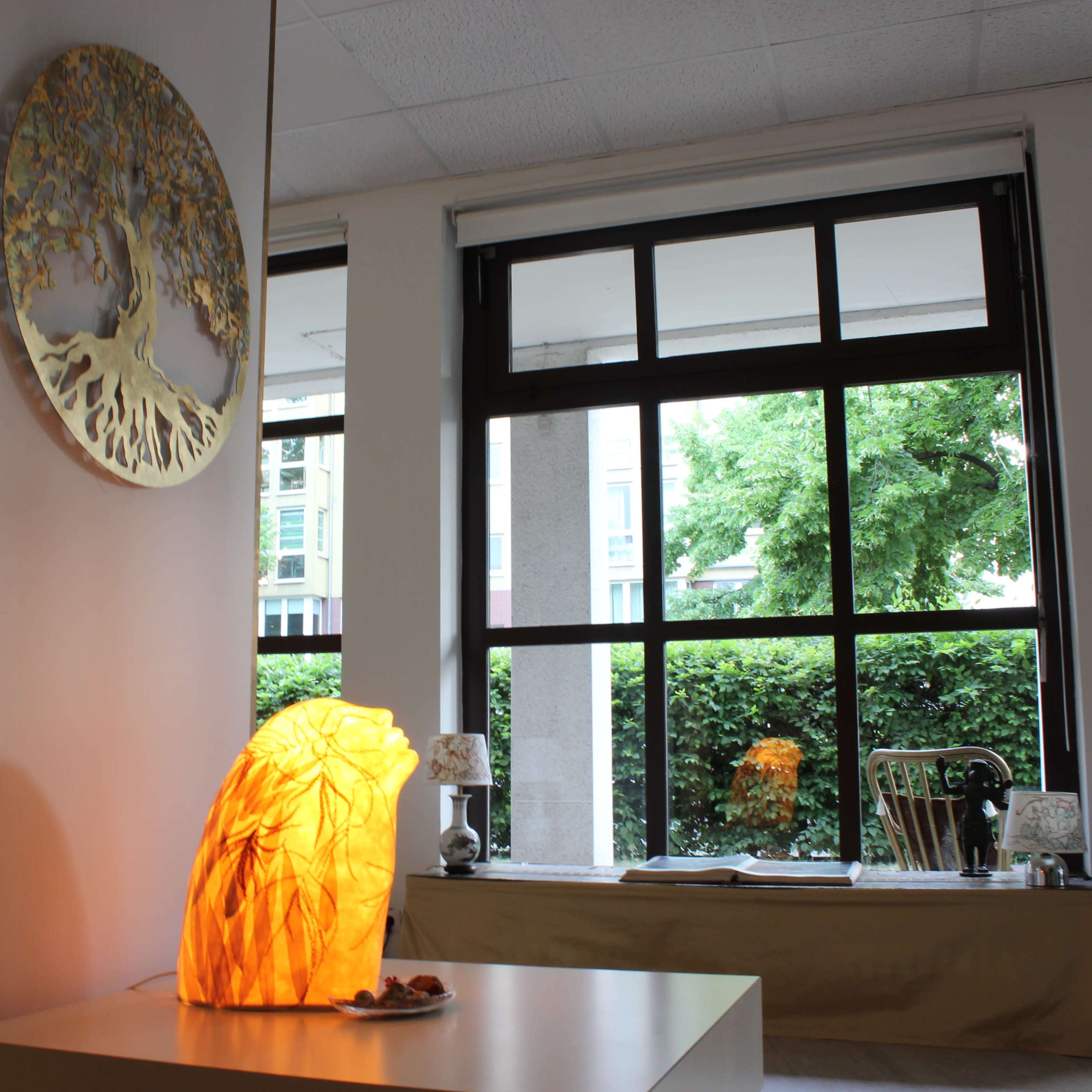 A glowing orange lamp is positioned on a white table in a room with large windows overlooking greenery.
