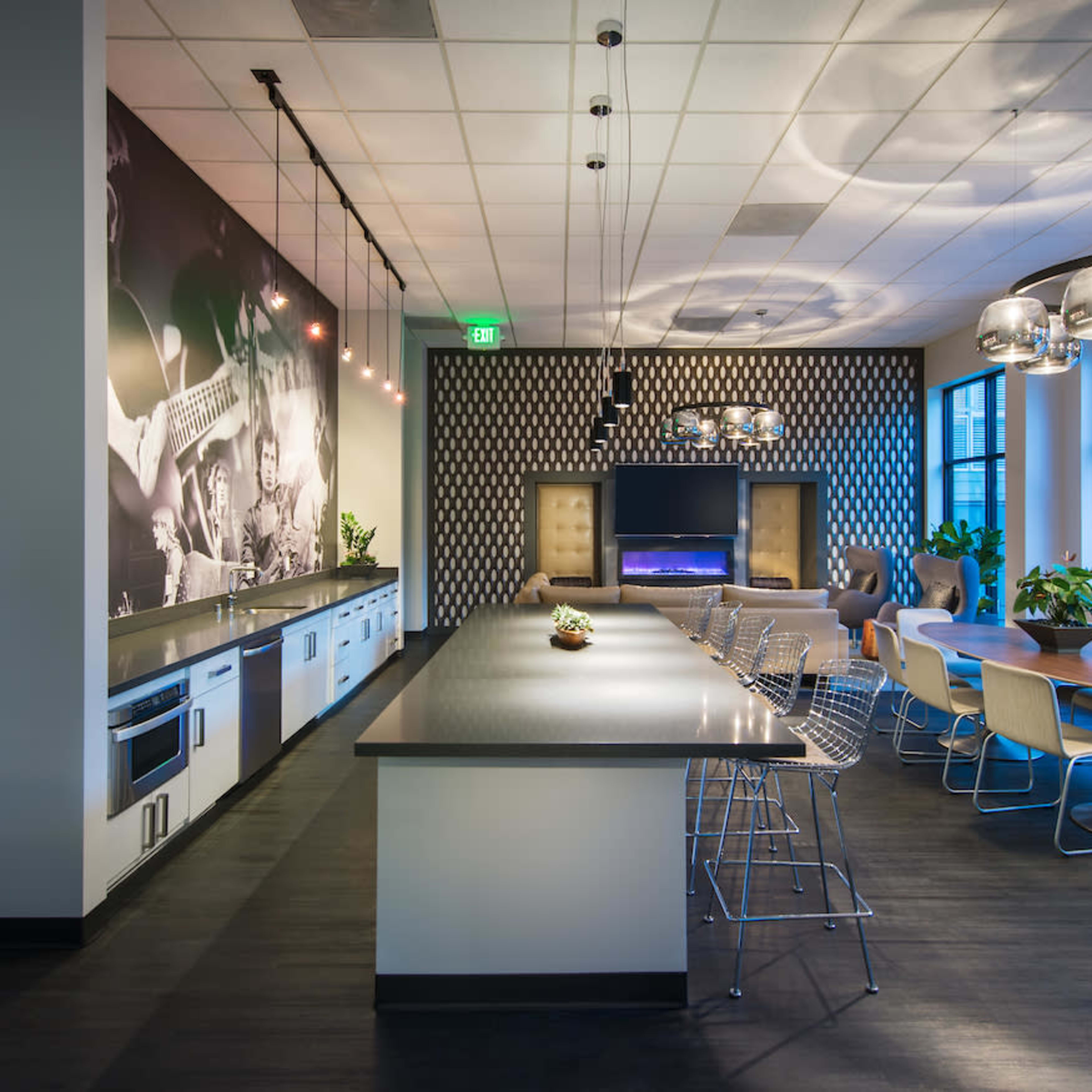 A modern communal kitchen and dining area with a long countertop, chairs, and a large television on a patterned wall.