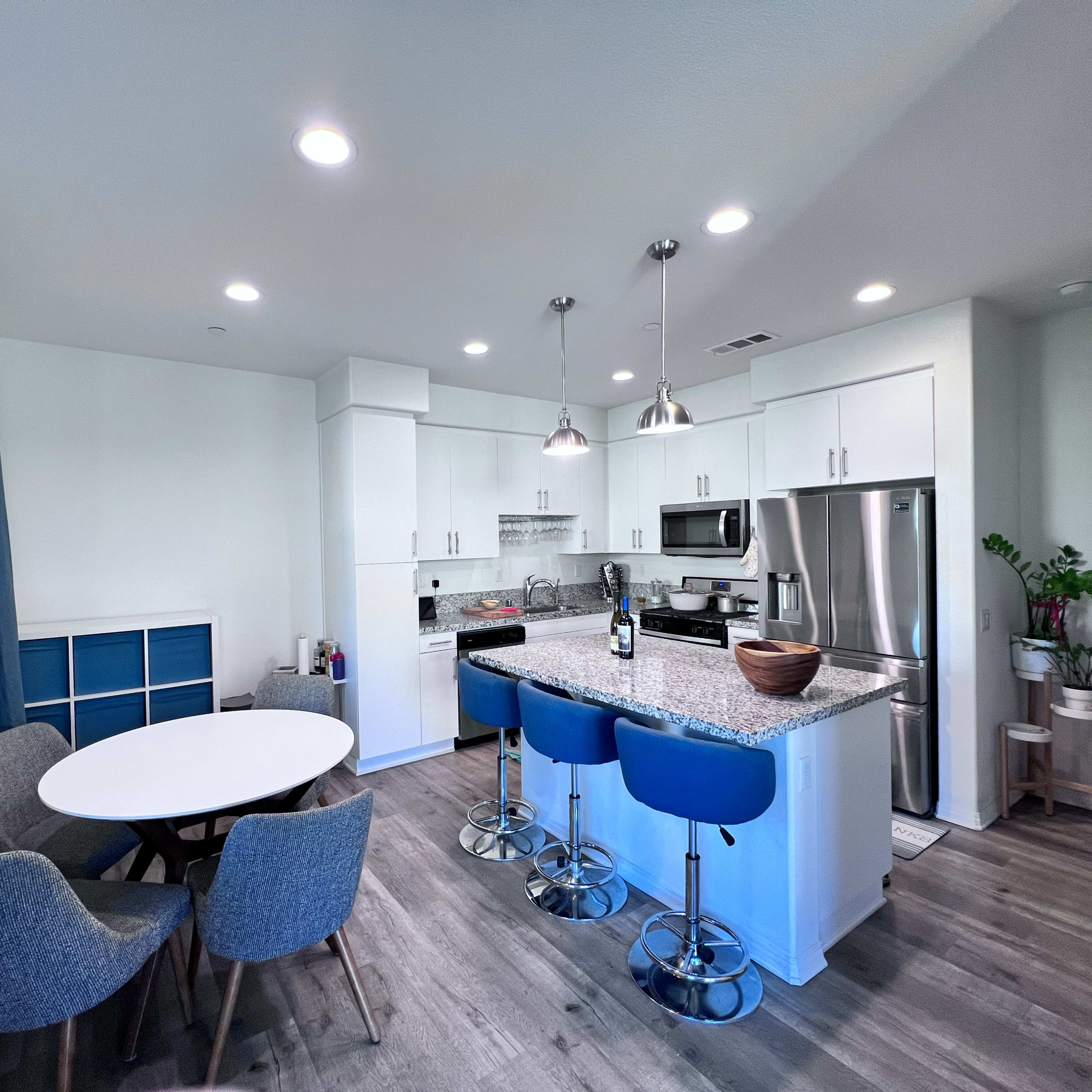 A modern kitchen features white cabinets, a gray countertop, stainless steel appliances, and a dining area with a round table and blue chairs.
