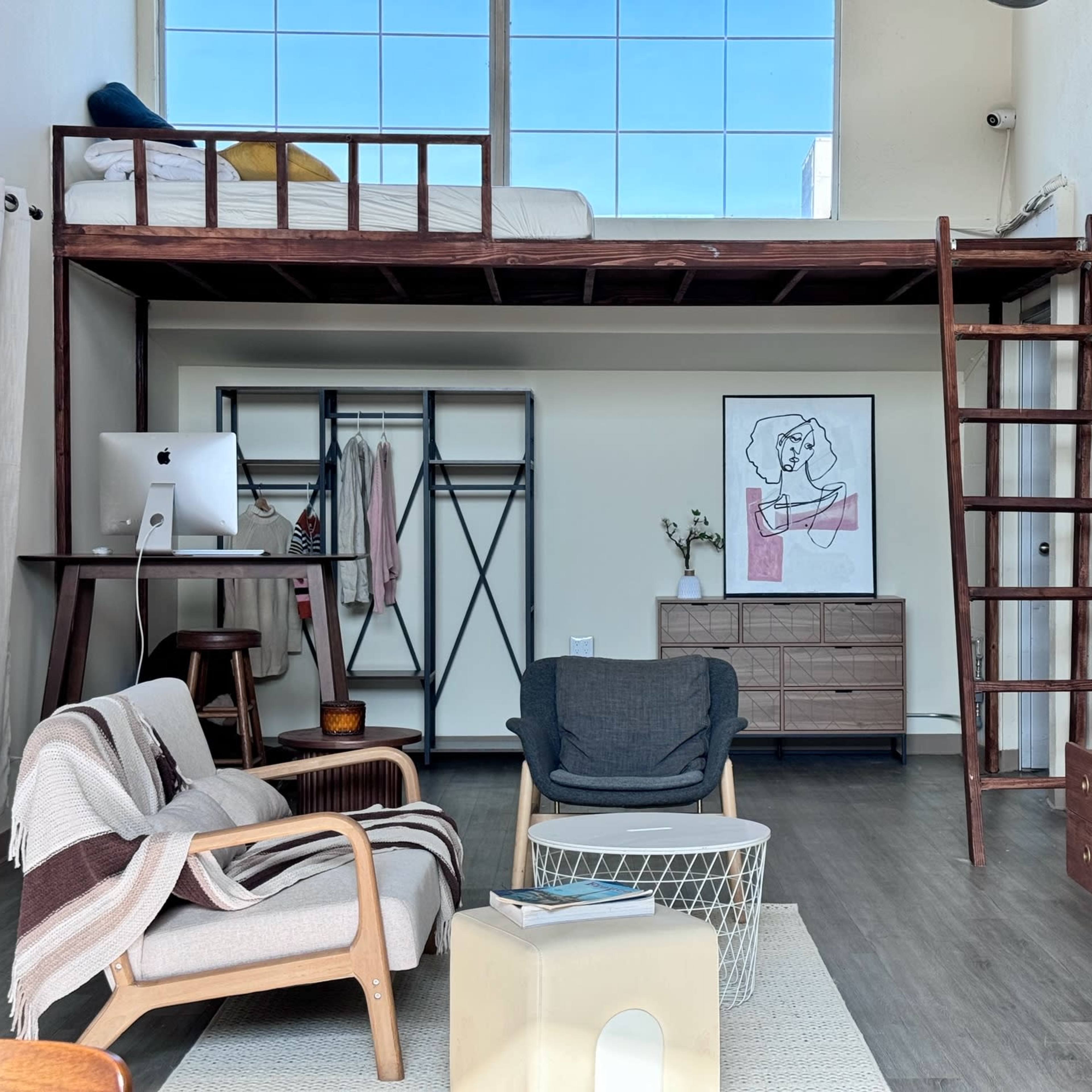 A lofted bed is positioned above a living area with a seating arrangement, storage furniture, and large windows allowing natural light.
