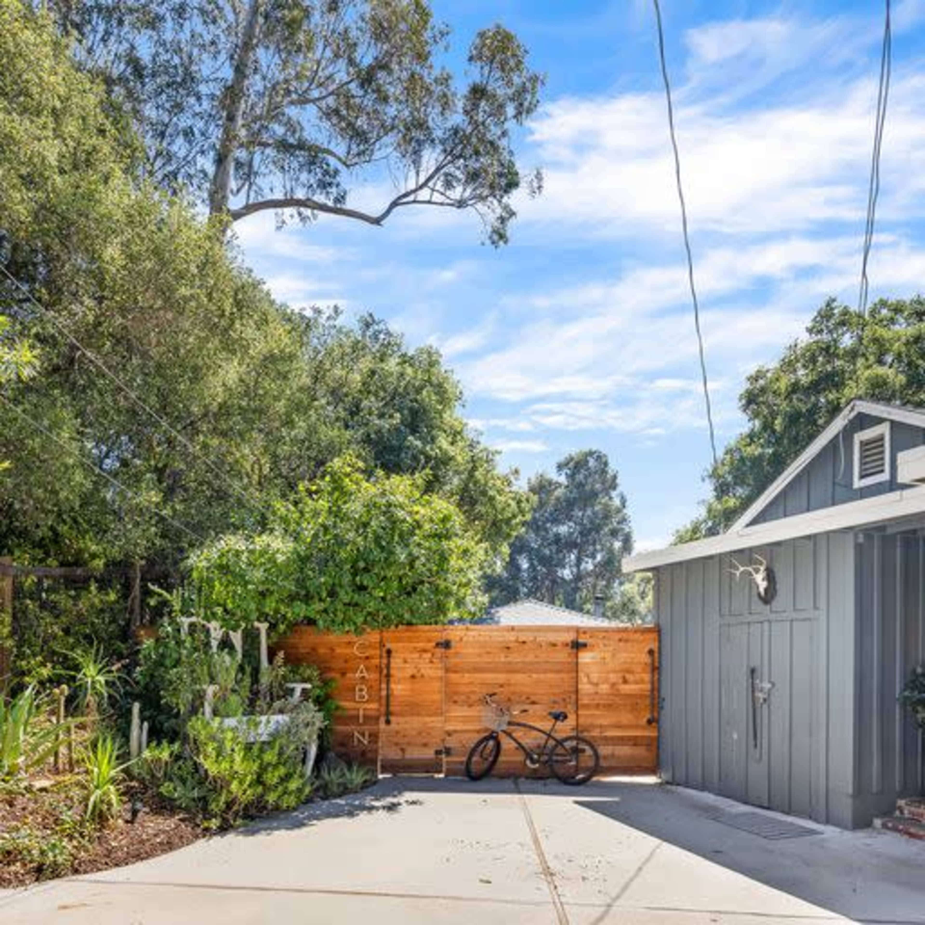 A gray cabin with a wooden fence and bicycles parked beside it is surrounded by lush greenery and trees.