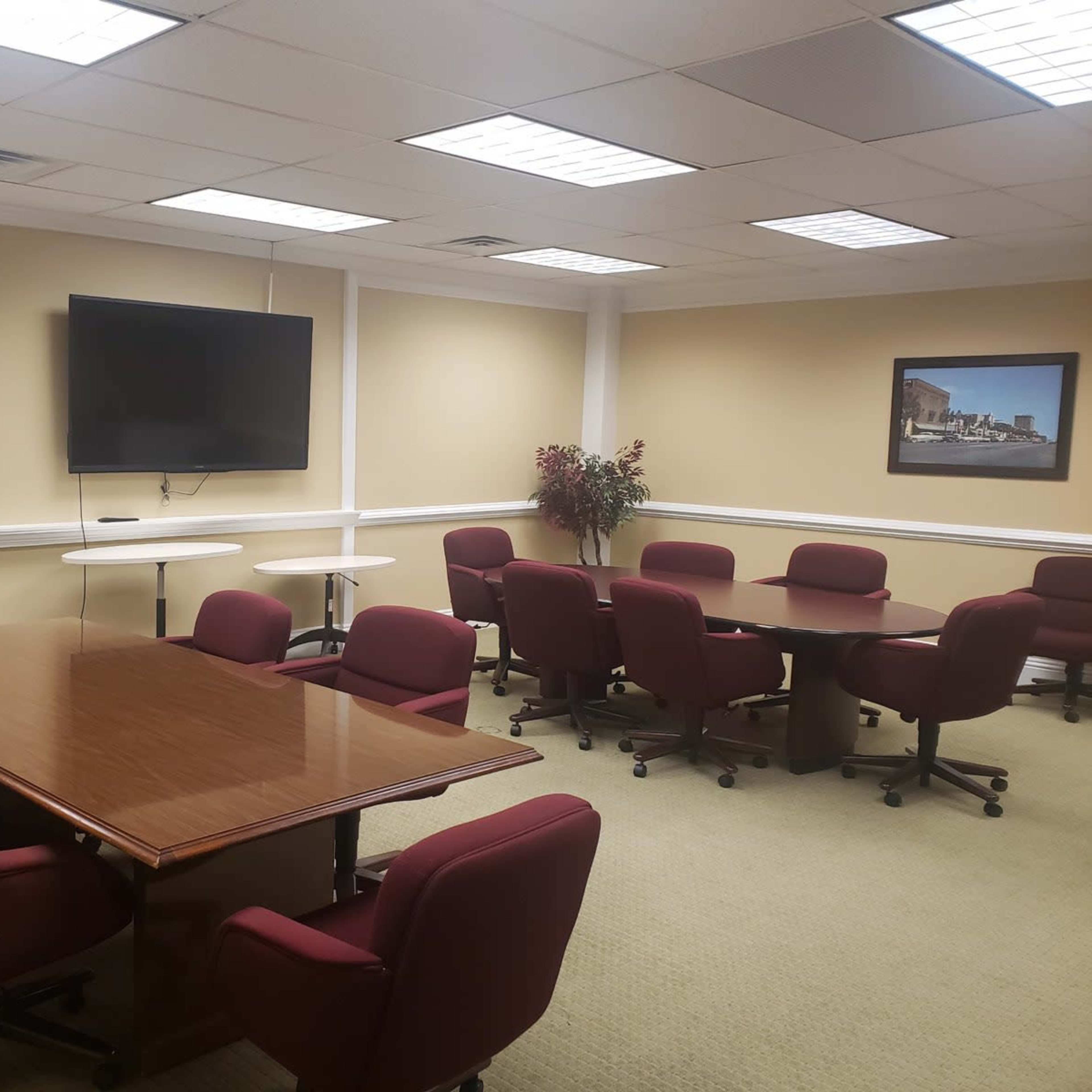 A conference room features several maroon chairs arranged around two tables and a wall-mounted television, with light-colored walls and a framed picture on the far wall.