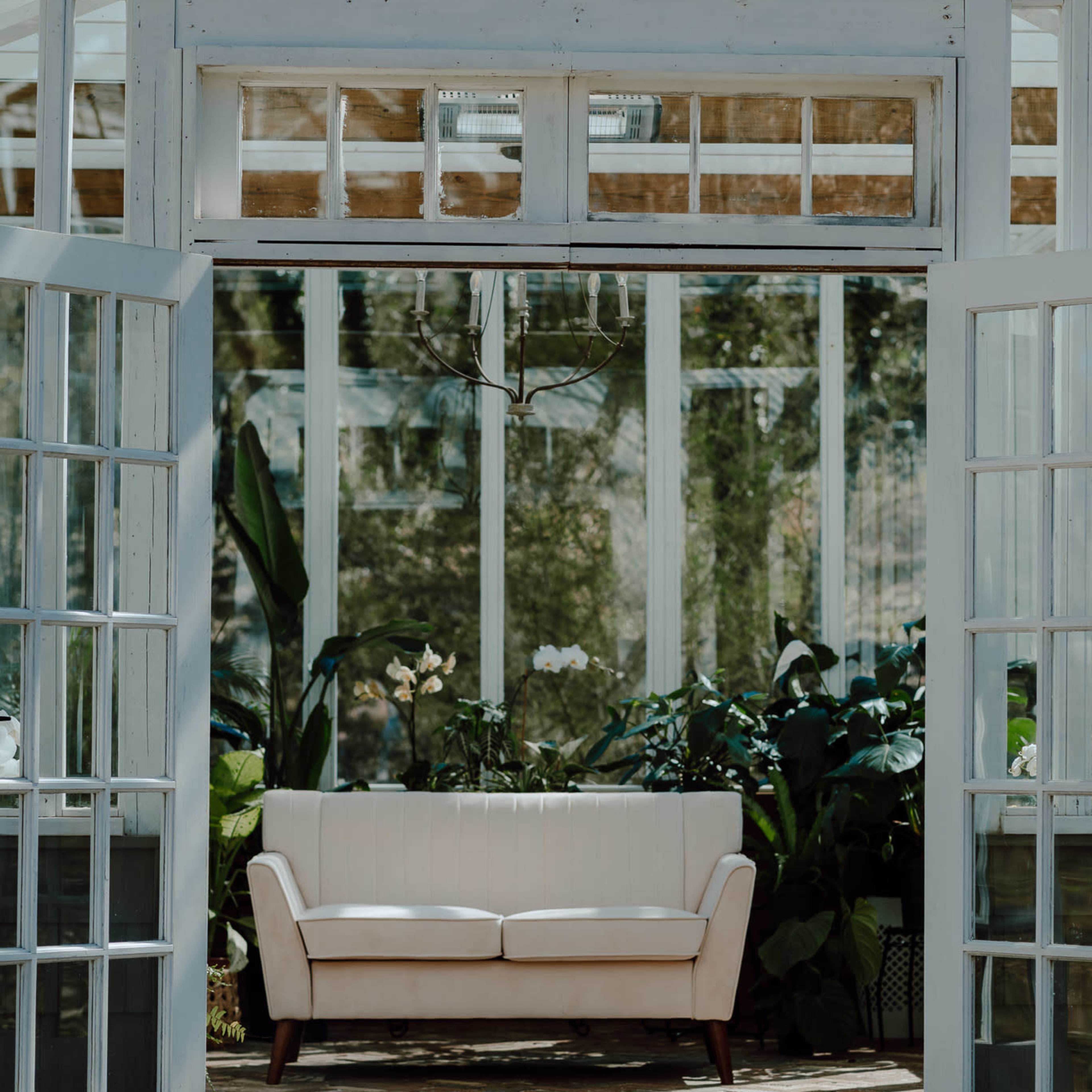 A white sofa sits inside a glass greenhouse surrounded by lush plants.