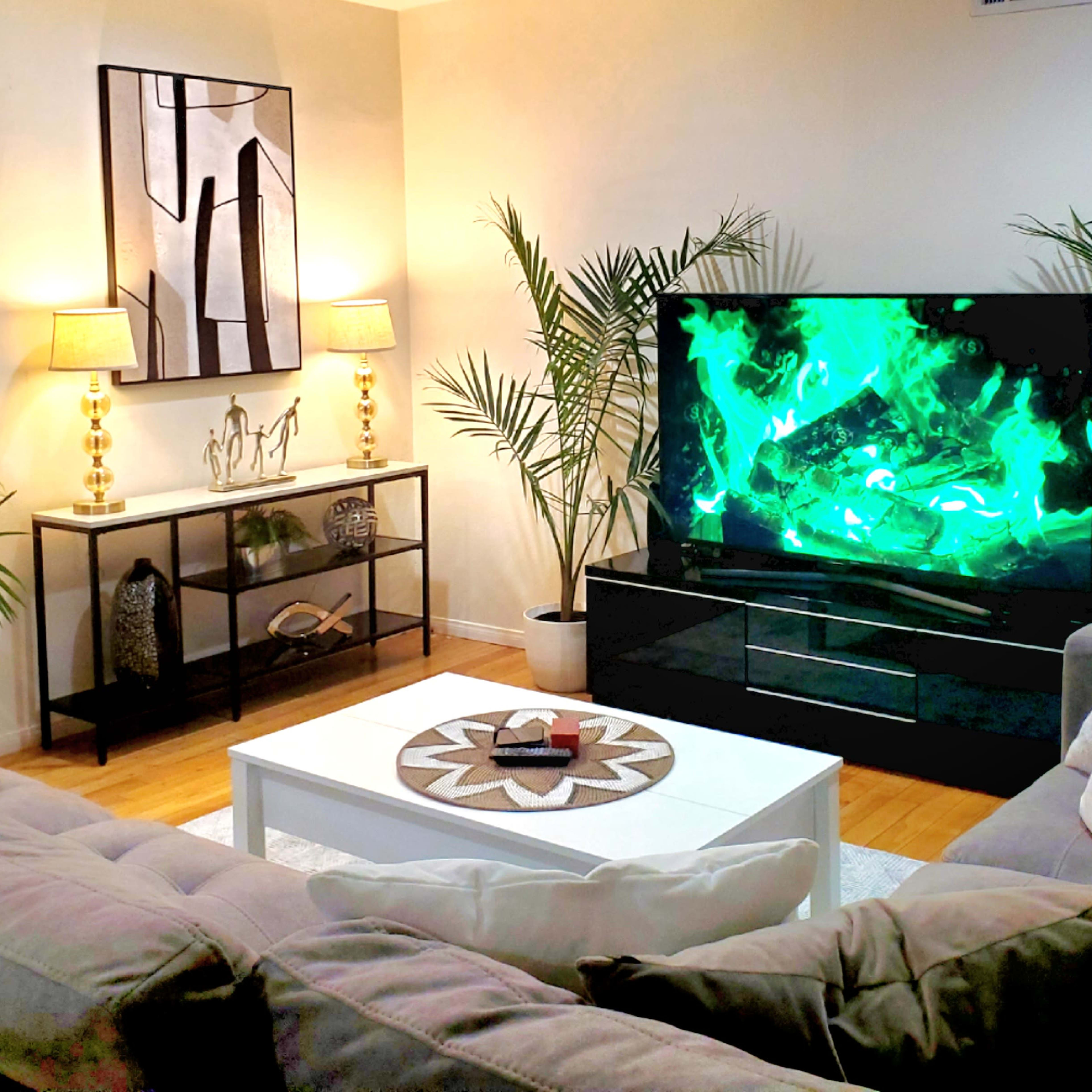 A modern living room with a large TV displaying green visuals, a white coffee table, two lamps, and decorative plants.