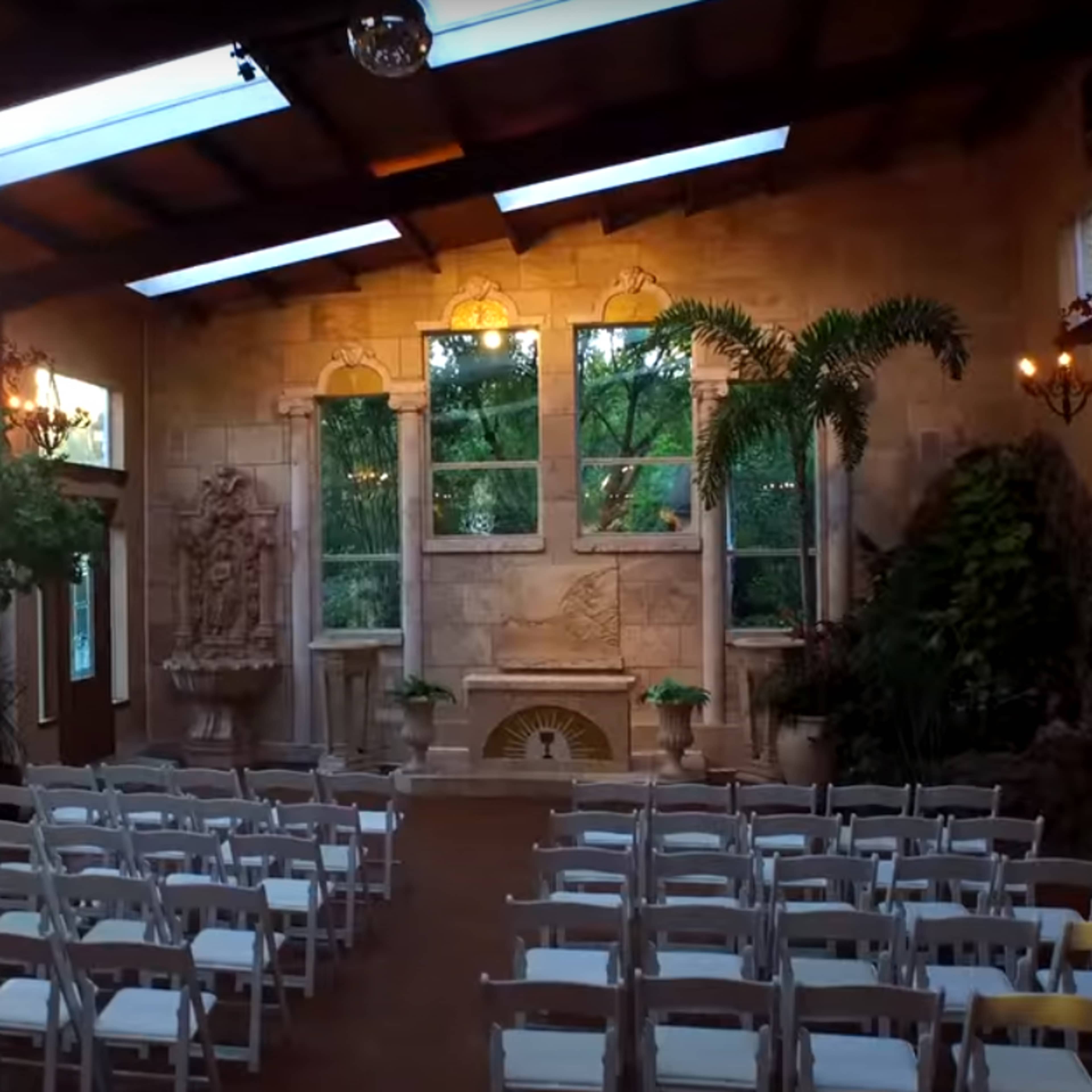 A spacious indoor venue set up for a ceremony, featuring rows of white chairs arranged facing a stone fireplace and large windows that allow natural light to enter.