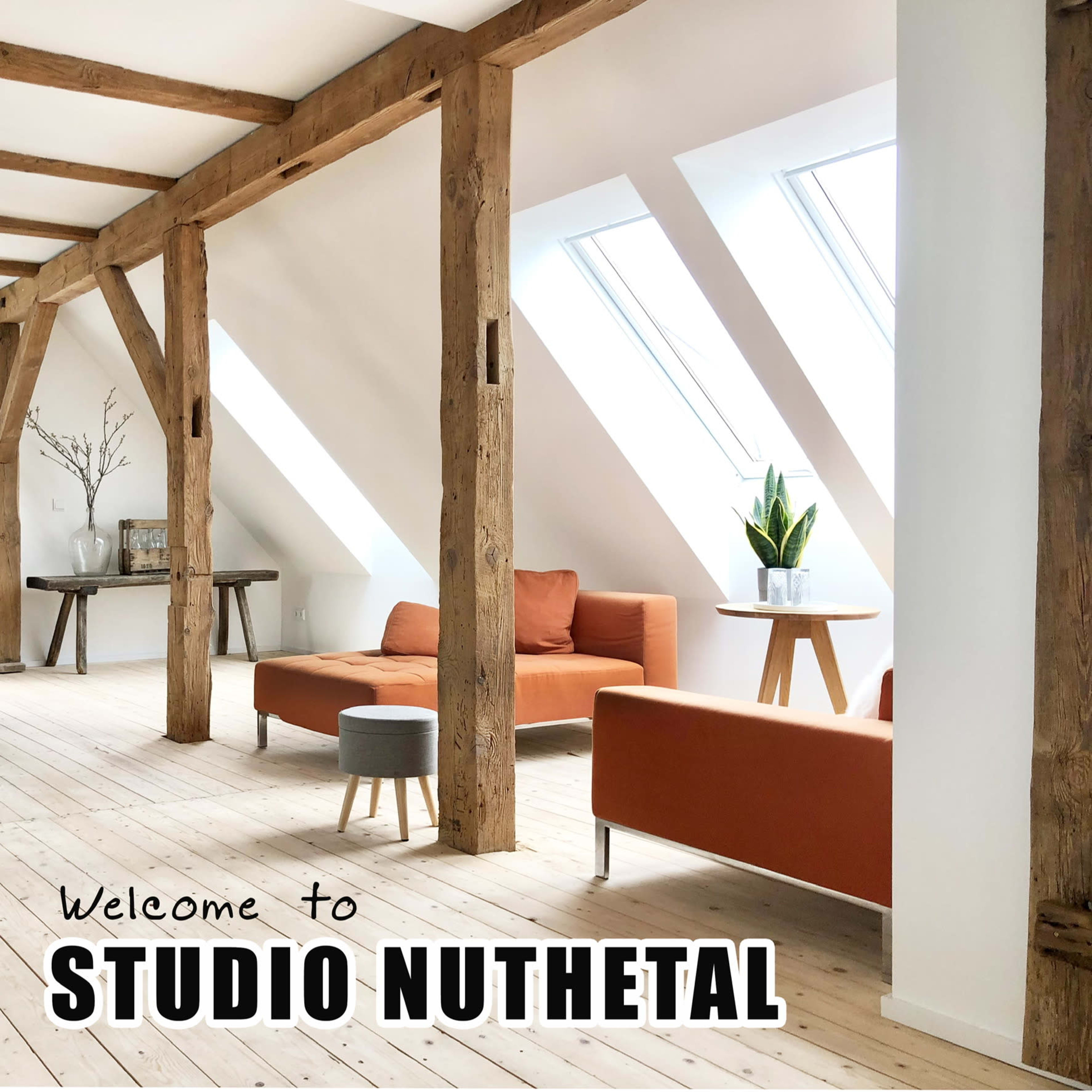 A bright, modern interior of a studio with wooden beams, orange couches, and large windows, featuring a small table with a plant.