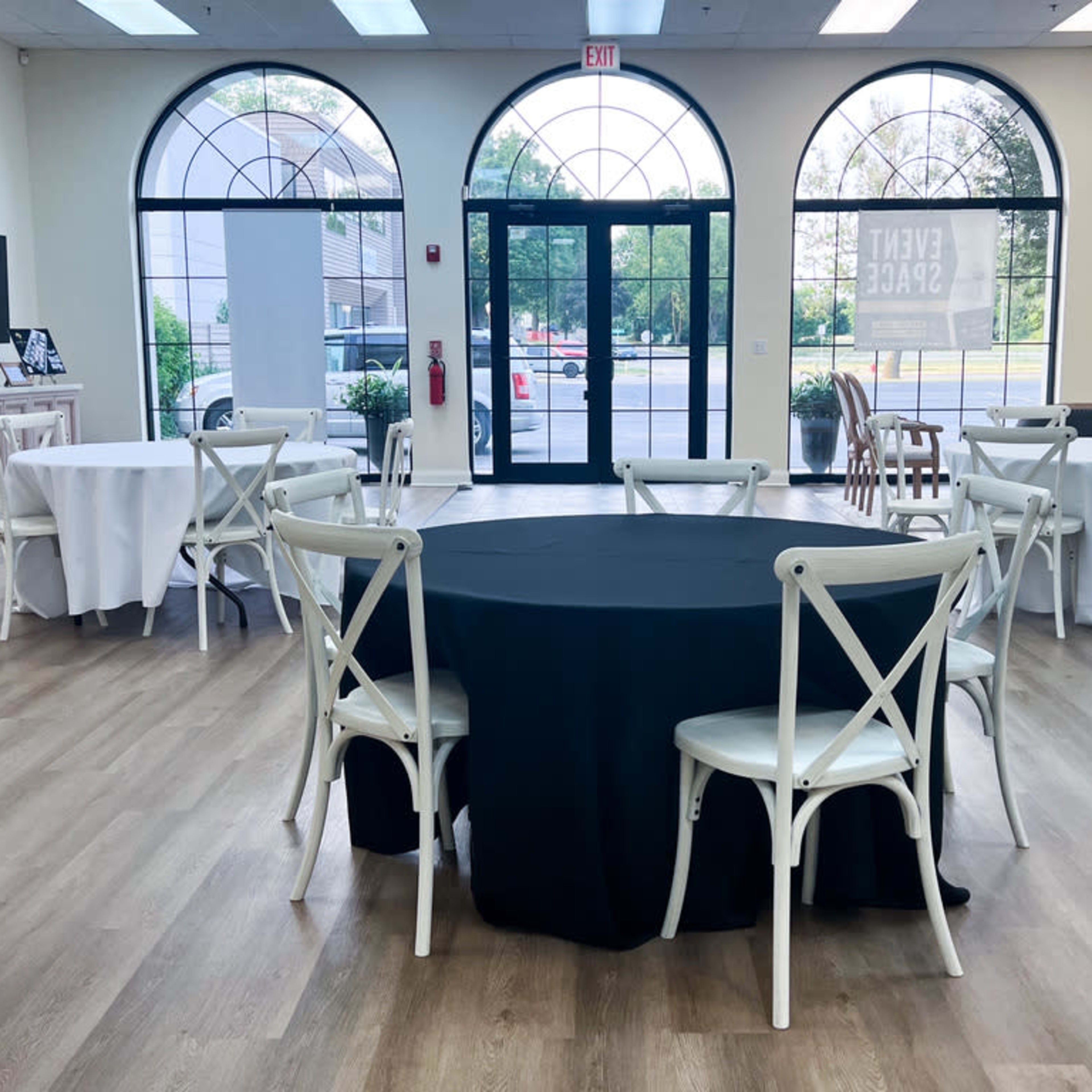 A spacious event room features several round tables with varying tablecloths and large windows allowing natural light.