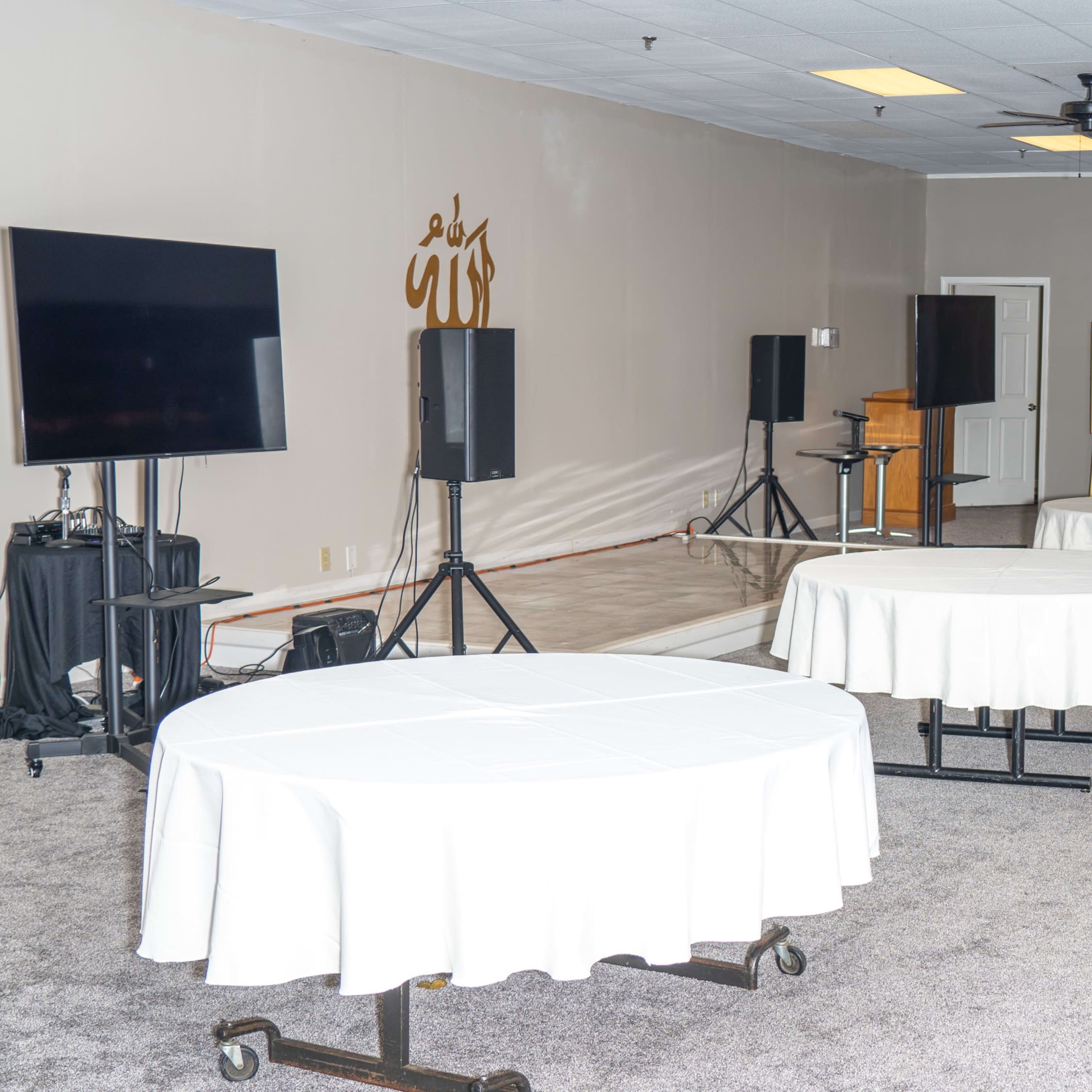 A room set up for an event with several round tables covered in white tablecloths, audio equipment, and a blank presentation board.