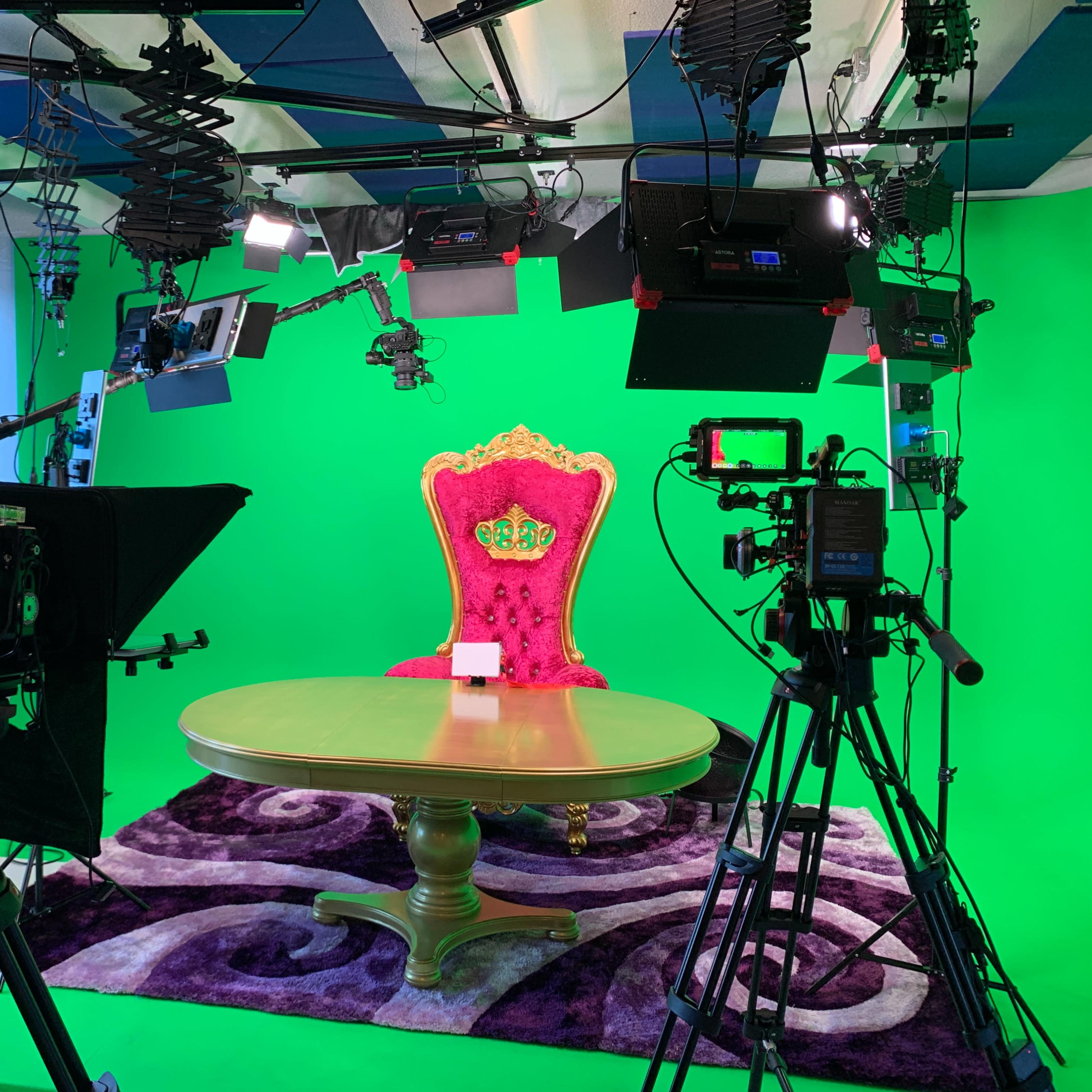 A studio setup featuring a large pink throne, a round table, and several cameras and lighting equipment arranged around a green screen backdrop.