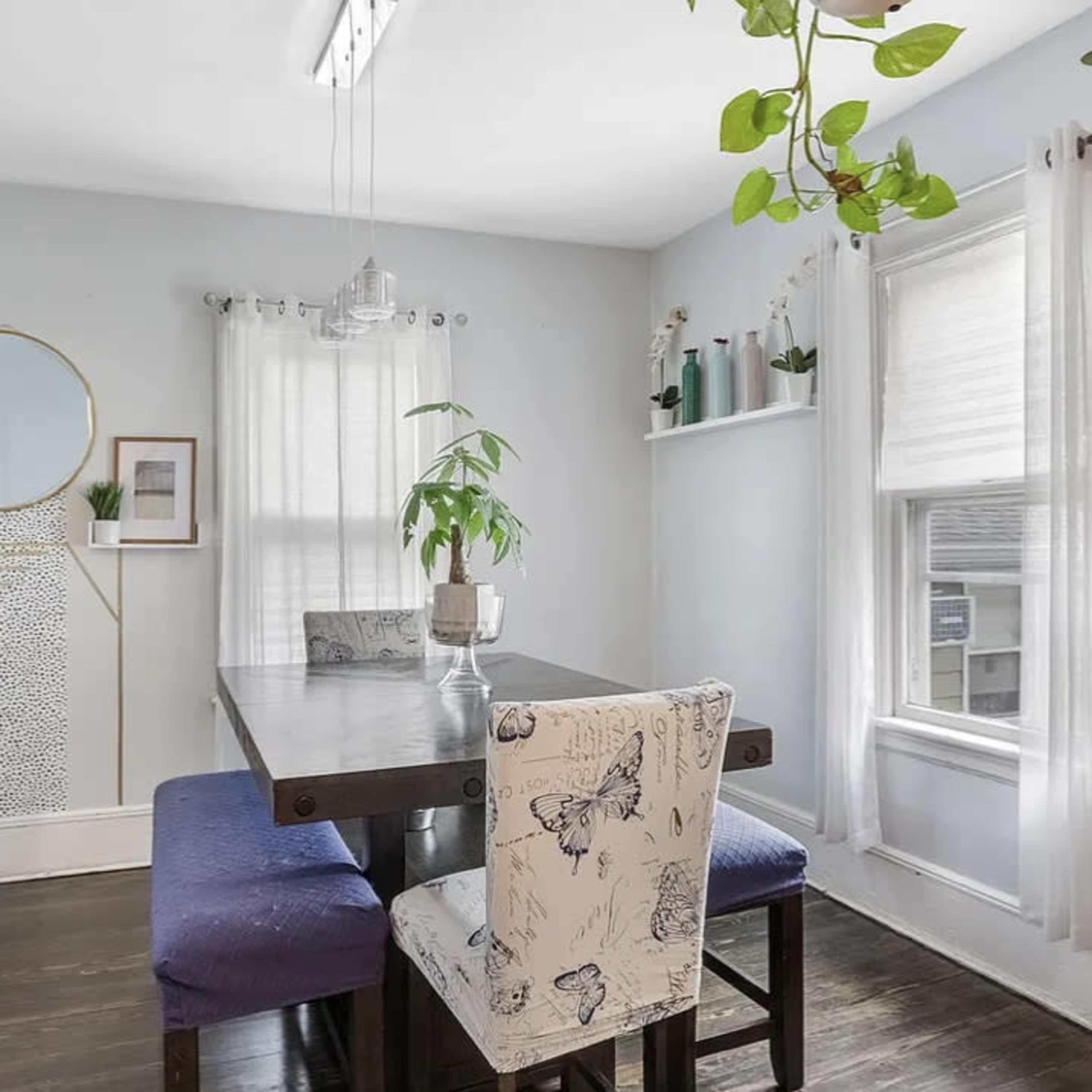 A dining area features a table with four chairs, plants decorating the space, and a large mirror on the wall.