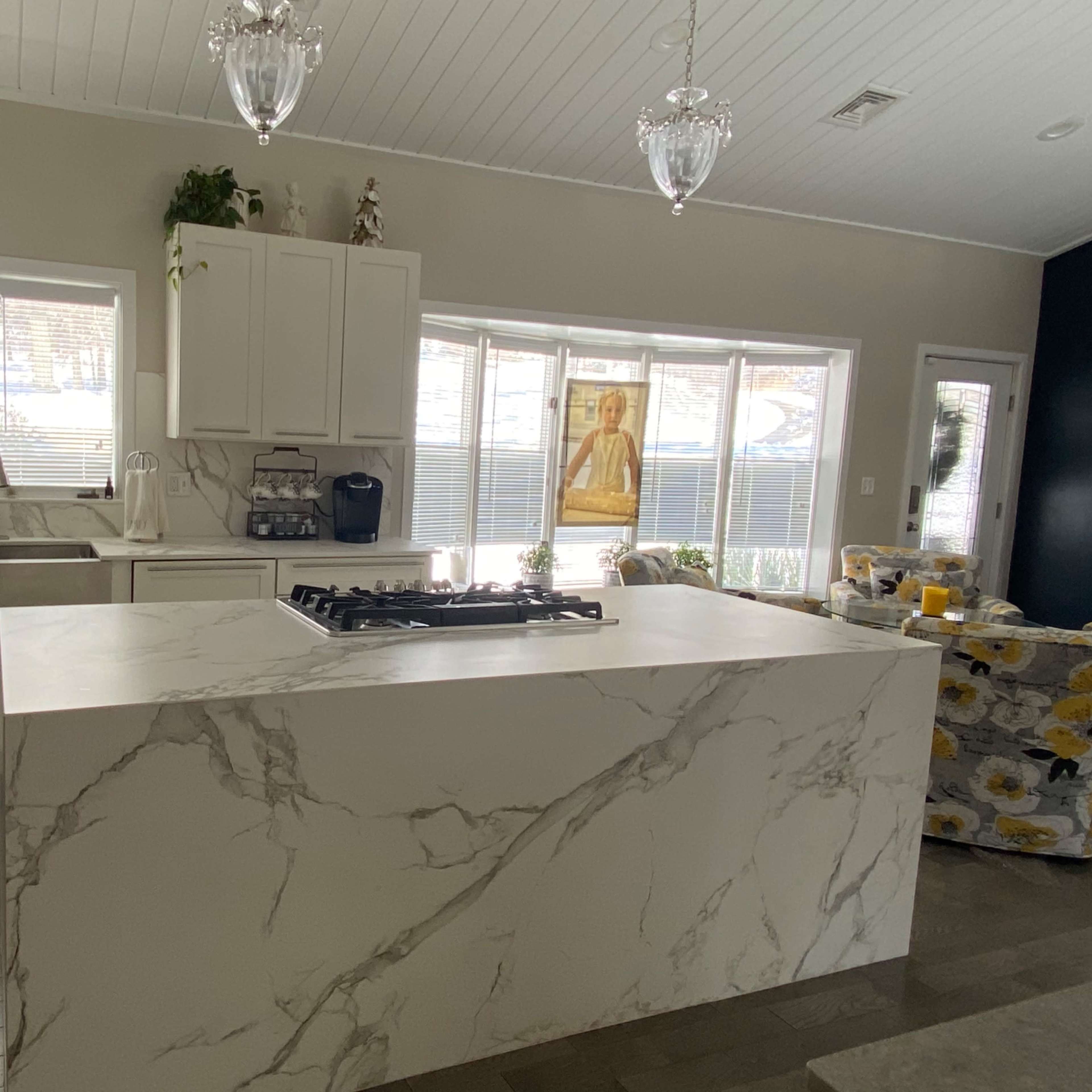 A modern kitchen features a large marble island with a gas cooktop, white cabinetry, and ample natural light from large windows.