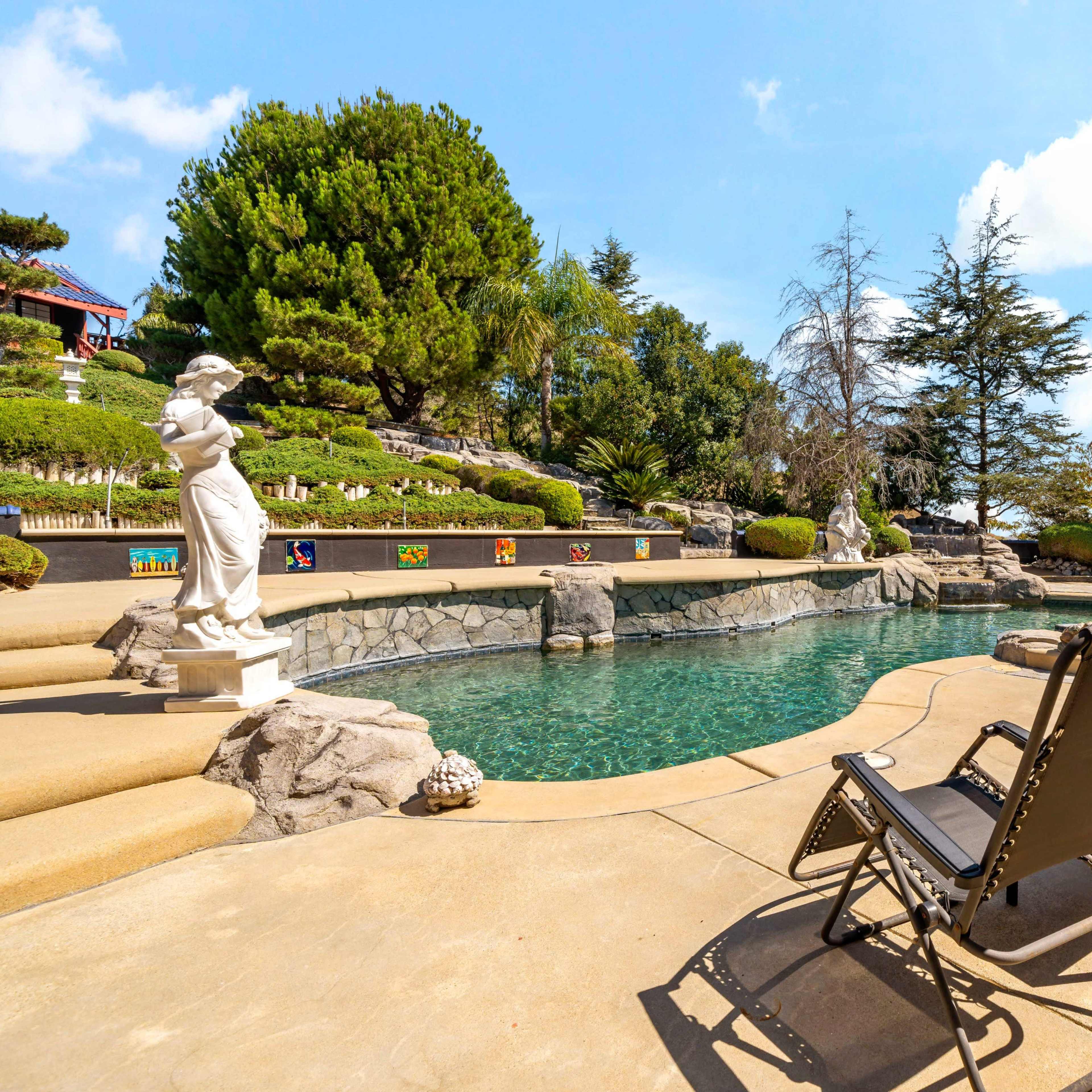 A landscaped backyard with a swimming pool, lounge chairs, and decorative statues surrounded by trees and shrubs.