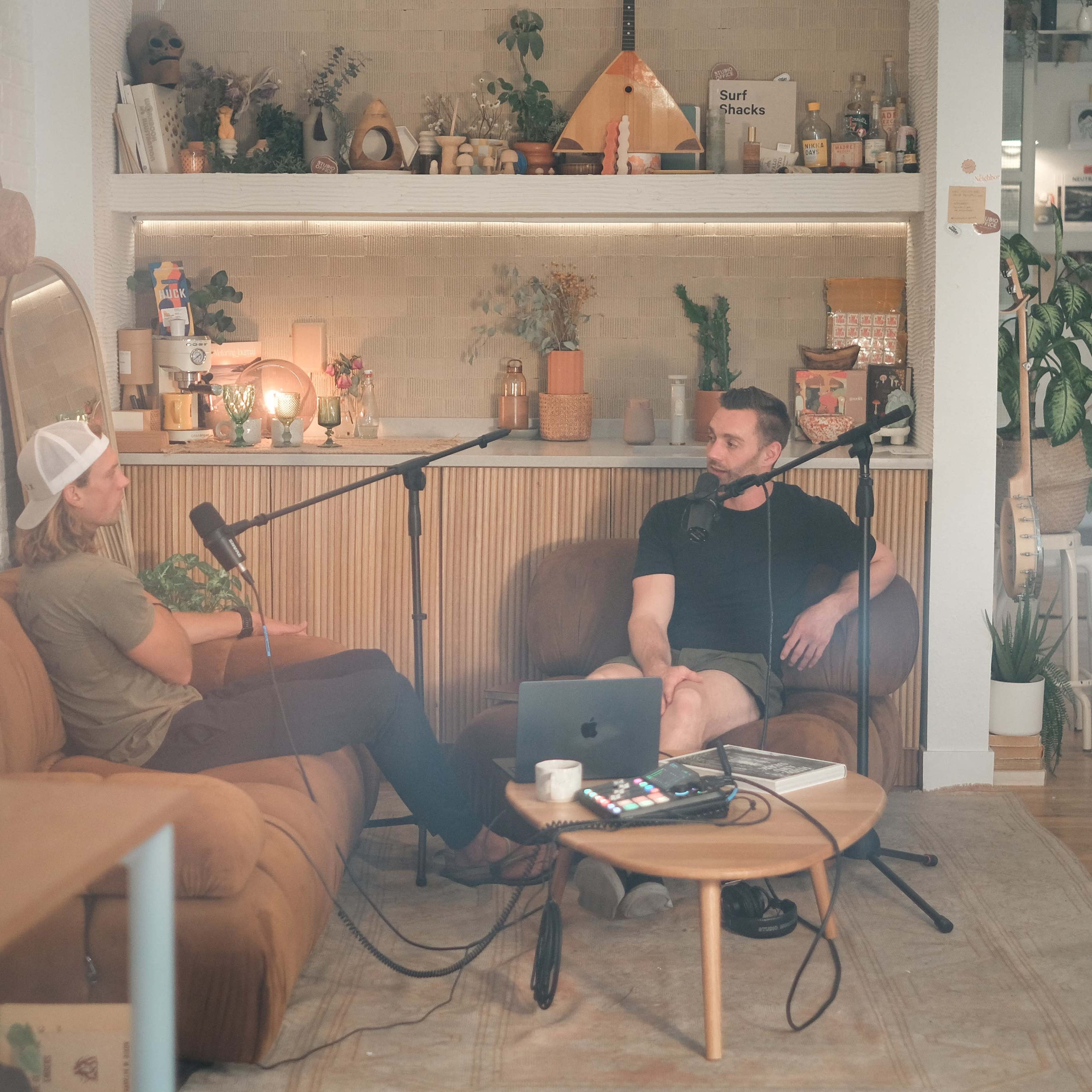 Two people are seated on a couch in a cozy studio, discussing into microphones while a laptop and various decorative items are visible around them.