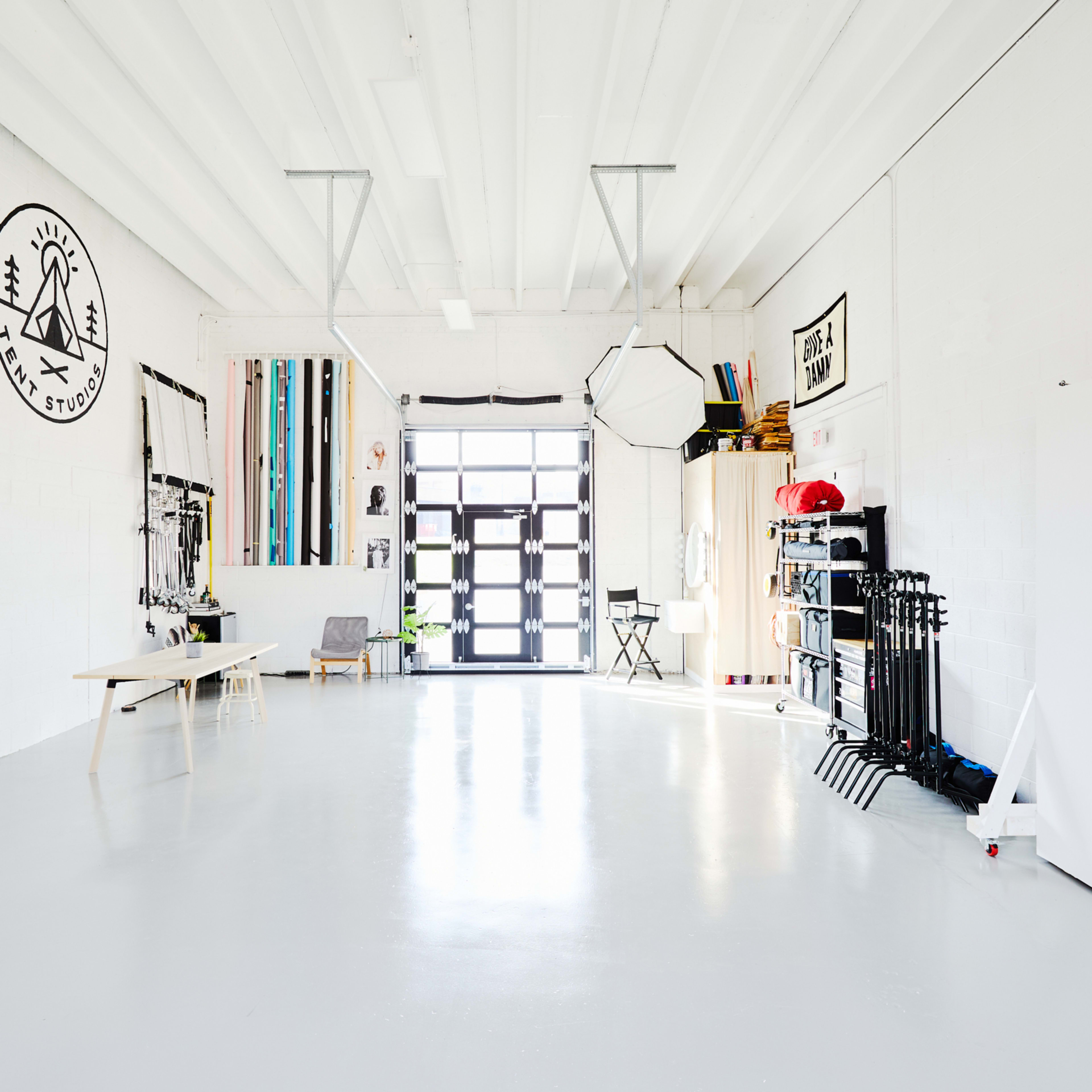 A spacious, bright studio with white walls, a large entrance door, and various photography equipment neatly organized along one side.