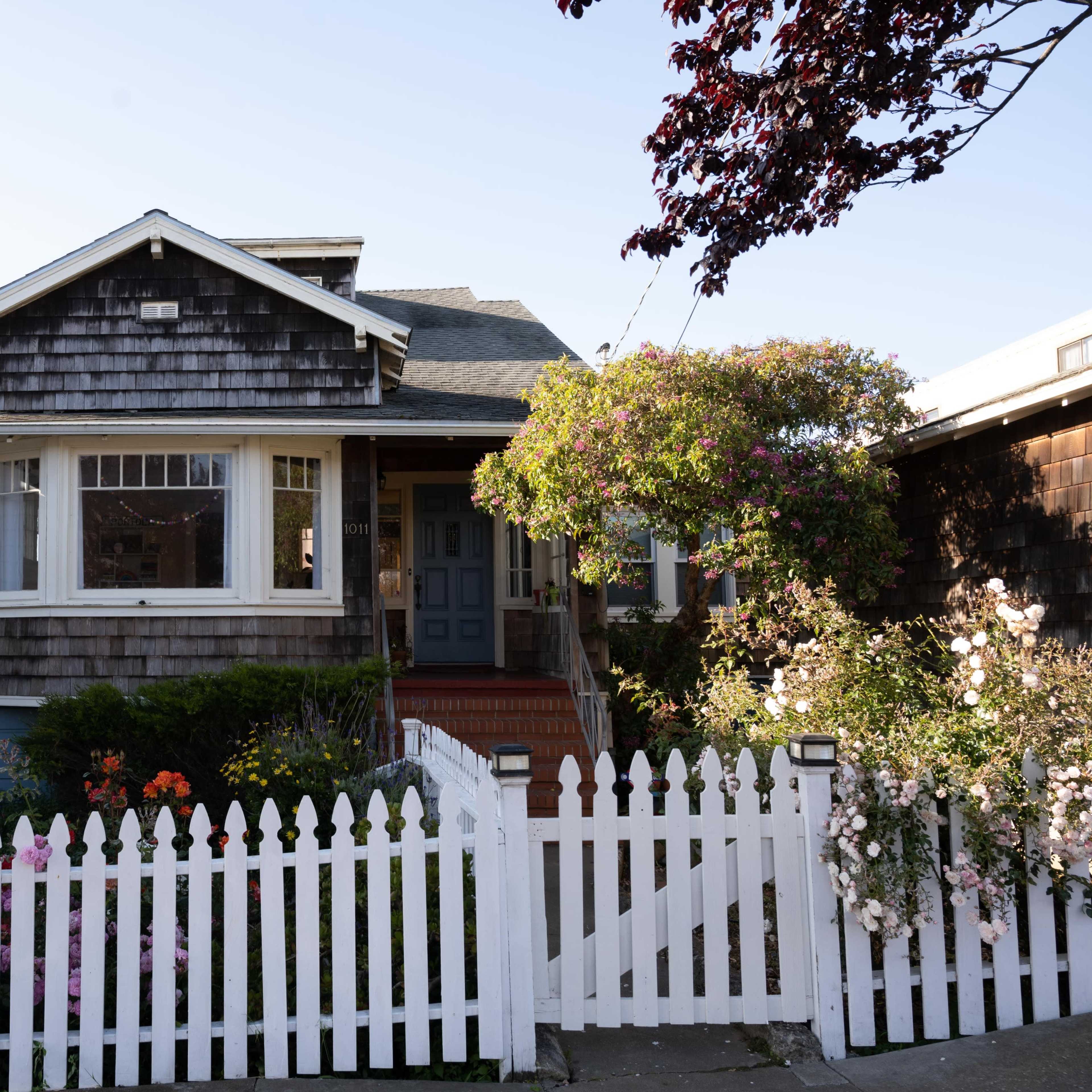 A wooden house with a shingled exterior and a front garden is enclosed by a white picket fence.