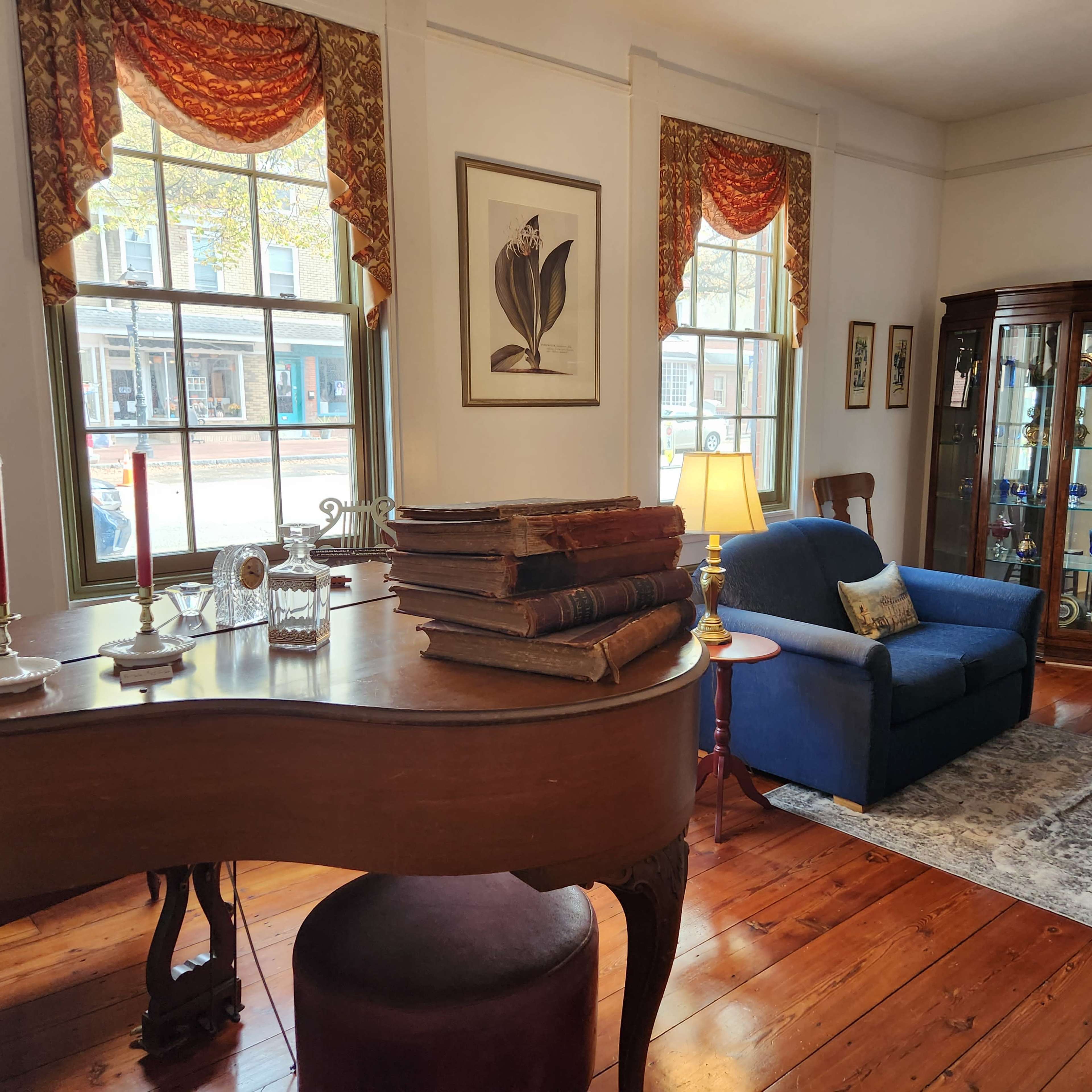 A well-furnished room featuring a piano, a blue armchair, a lamp, and shelves displaying decorative items and books near large windows with patterned curtains.