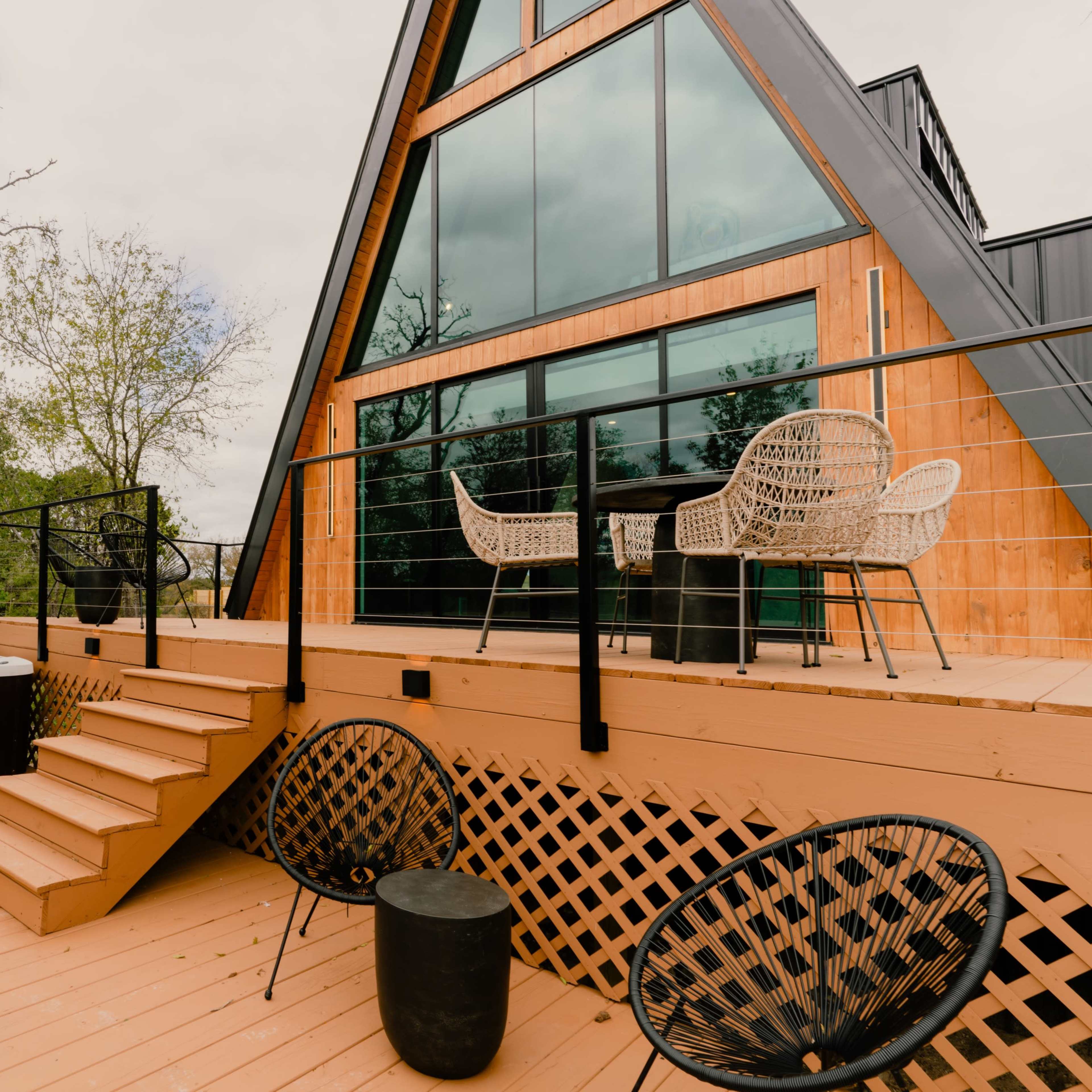 A modern A-frame house with large windows, a hot tub, and a wooden deck surrounded by trees.
