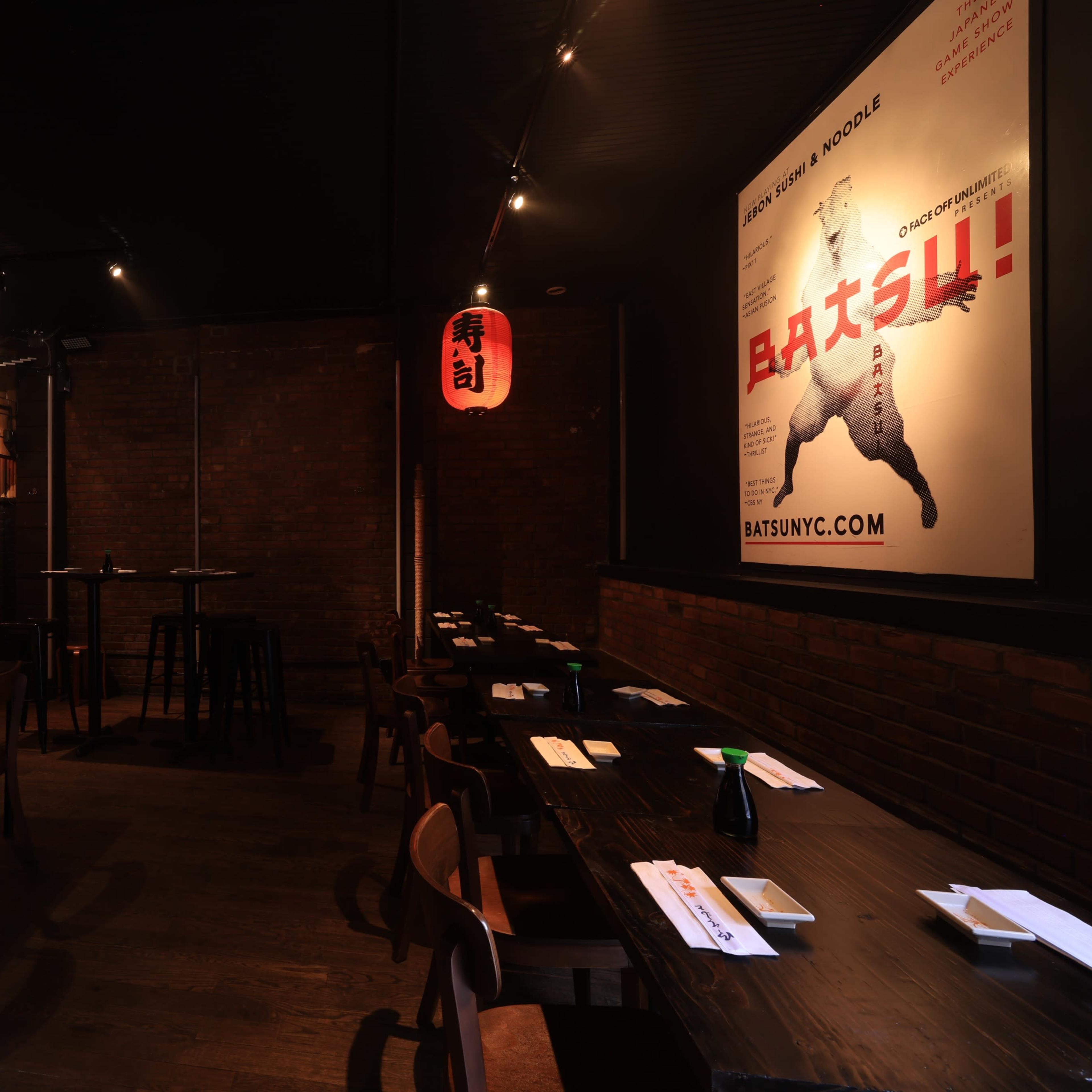 An empty restaurant interior with dark wooden tables, chairs, and red lanterns, featuring a large poster on the wall.