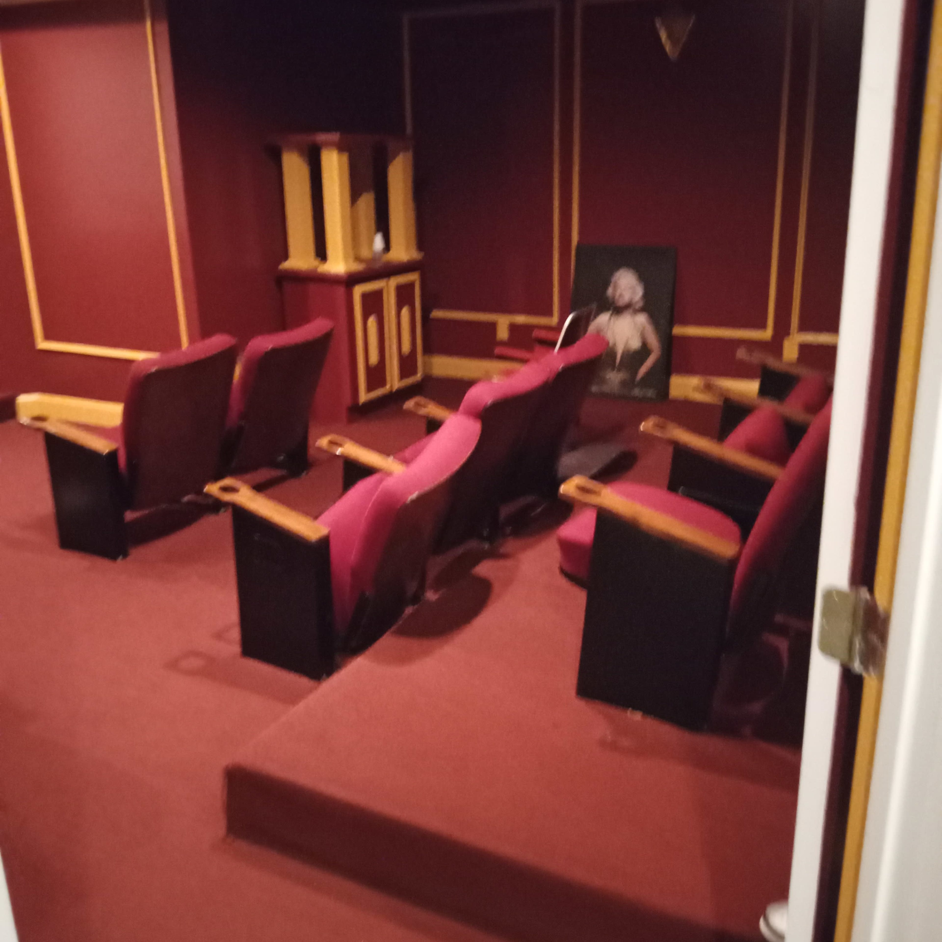 A small, dimly lit home theater with red walls and carpet, featuring several plush red seating chairs arranged in a semi-circle.