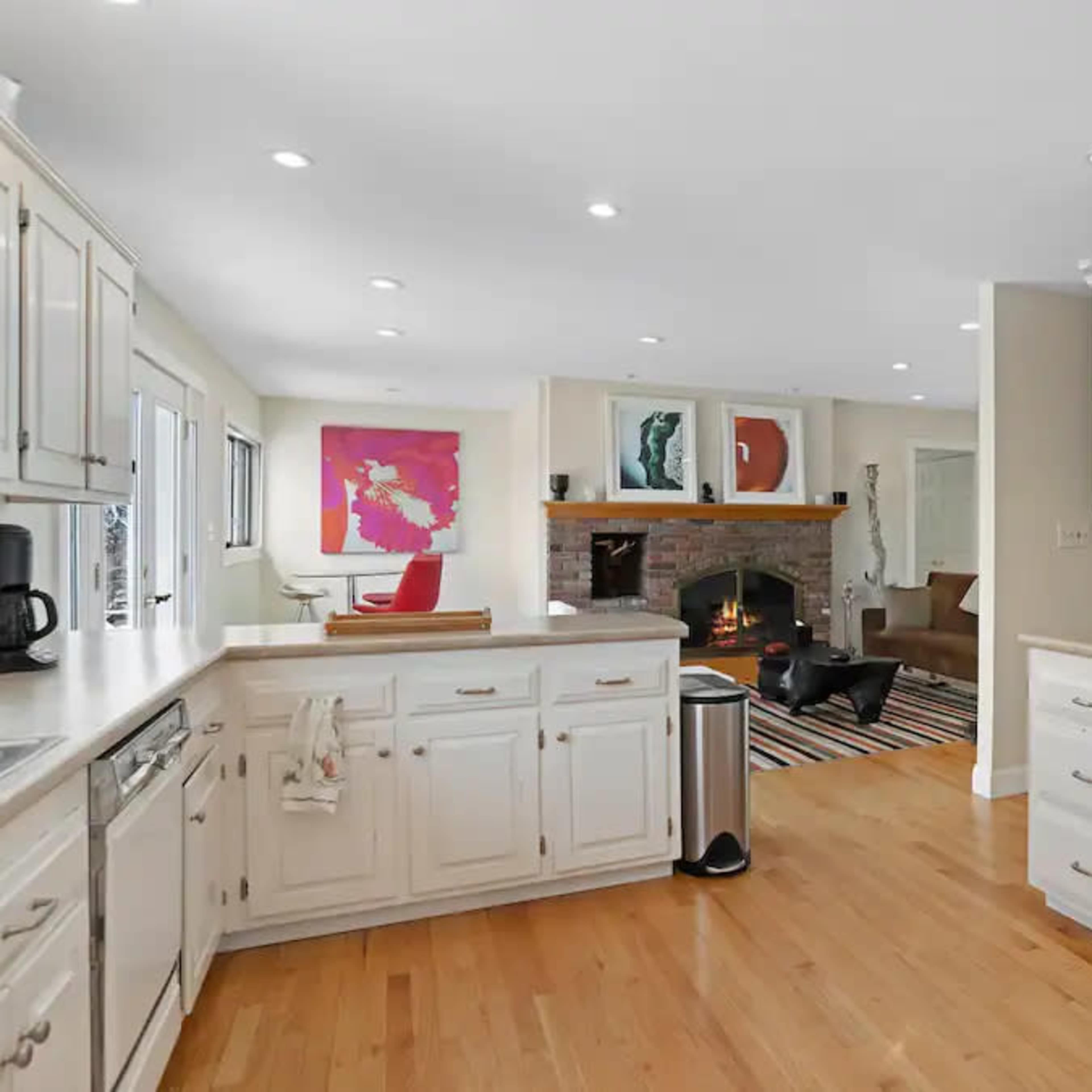 A modern kitchen features white cabinetry, a double sink, and an open layout that connects to a living area with a fireplace.
