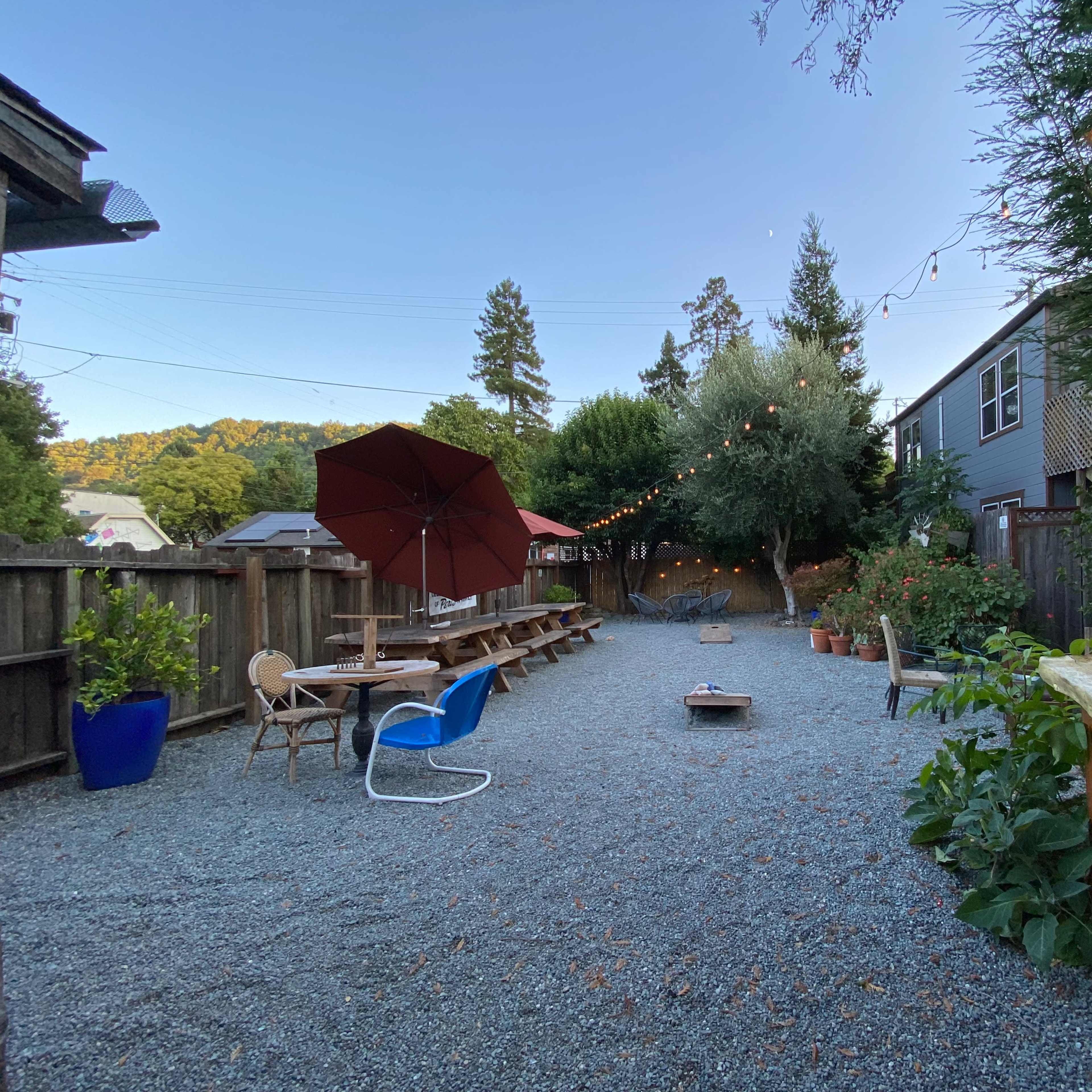 A gravel outdoor space with picnic tables, a large red umbrella, a fire pit, and greenery, surrounded by wooden fences and residential buildings.