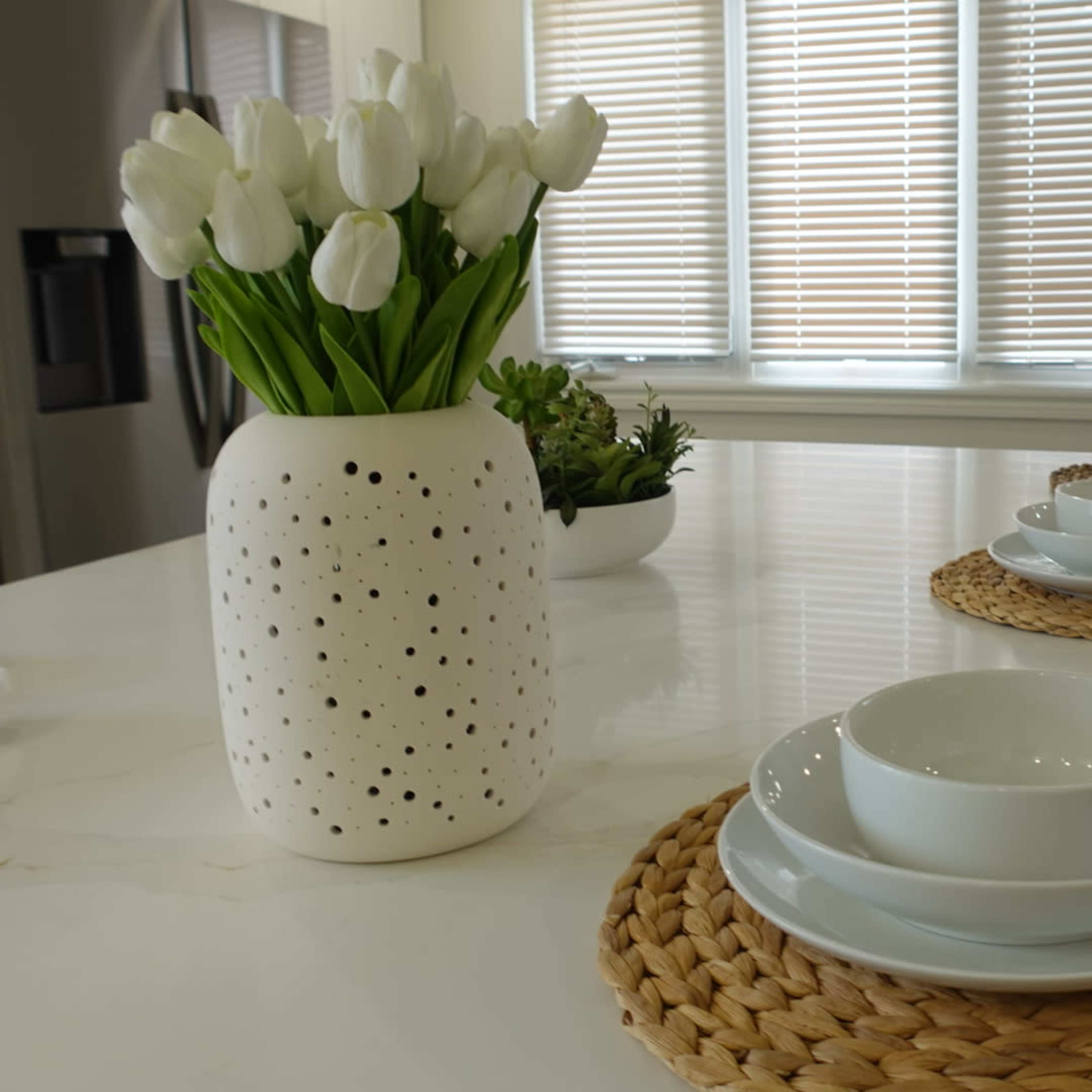 A dining area features a table set with white bowls on woven placemats, accompanied by a vase of tulips and a small plant in the background.