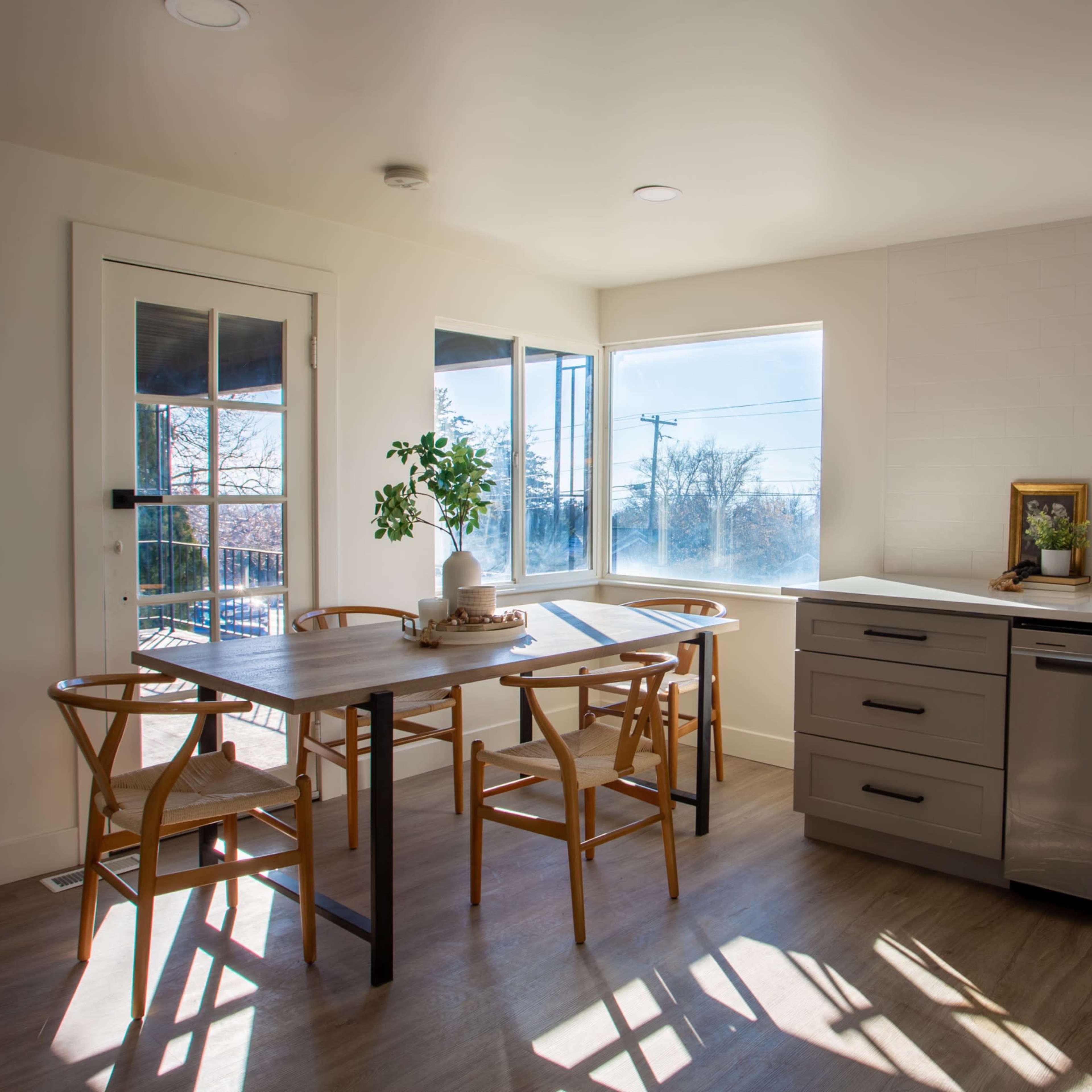 A modern kitchen features a wooden table with chairs, a sink, and large windows allowing natural light to fill the room.