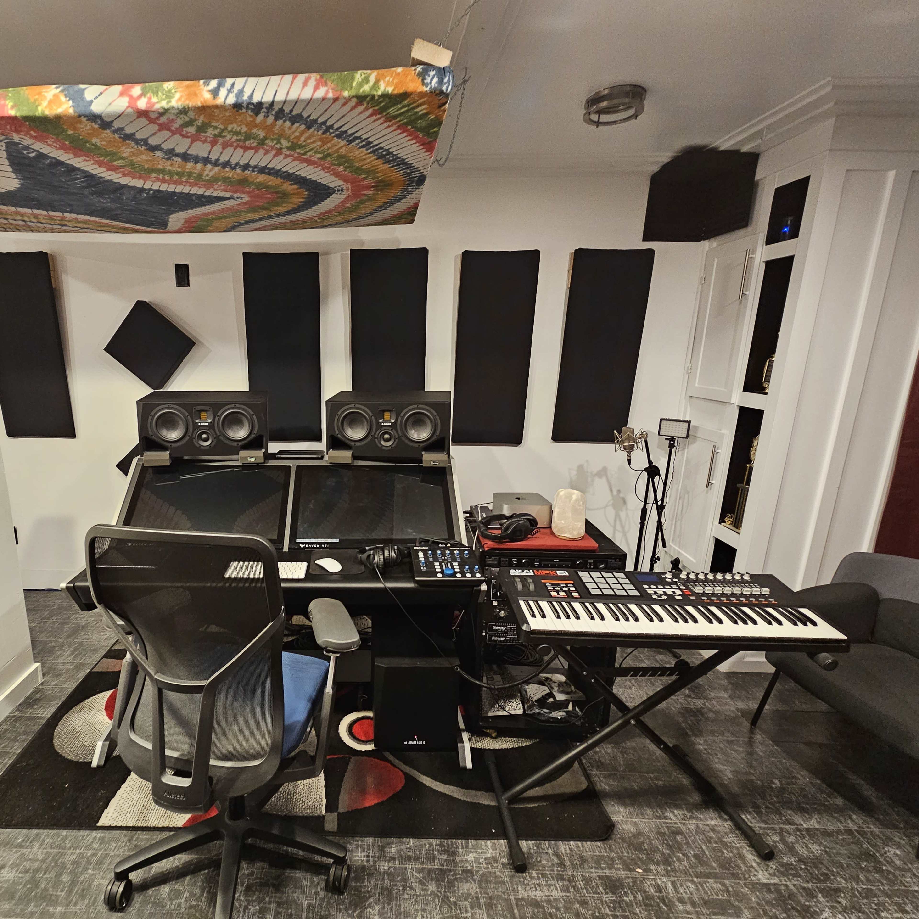 A music studio setup with a large desk featuring speakers, a mixer, a keyboard, and soundproofing panels on the walls.