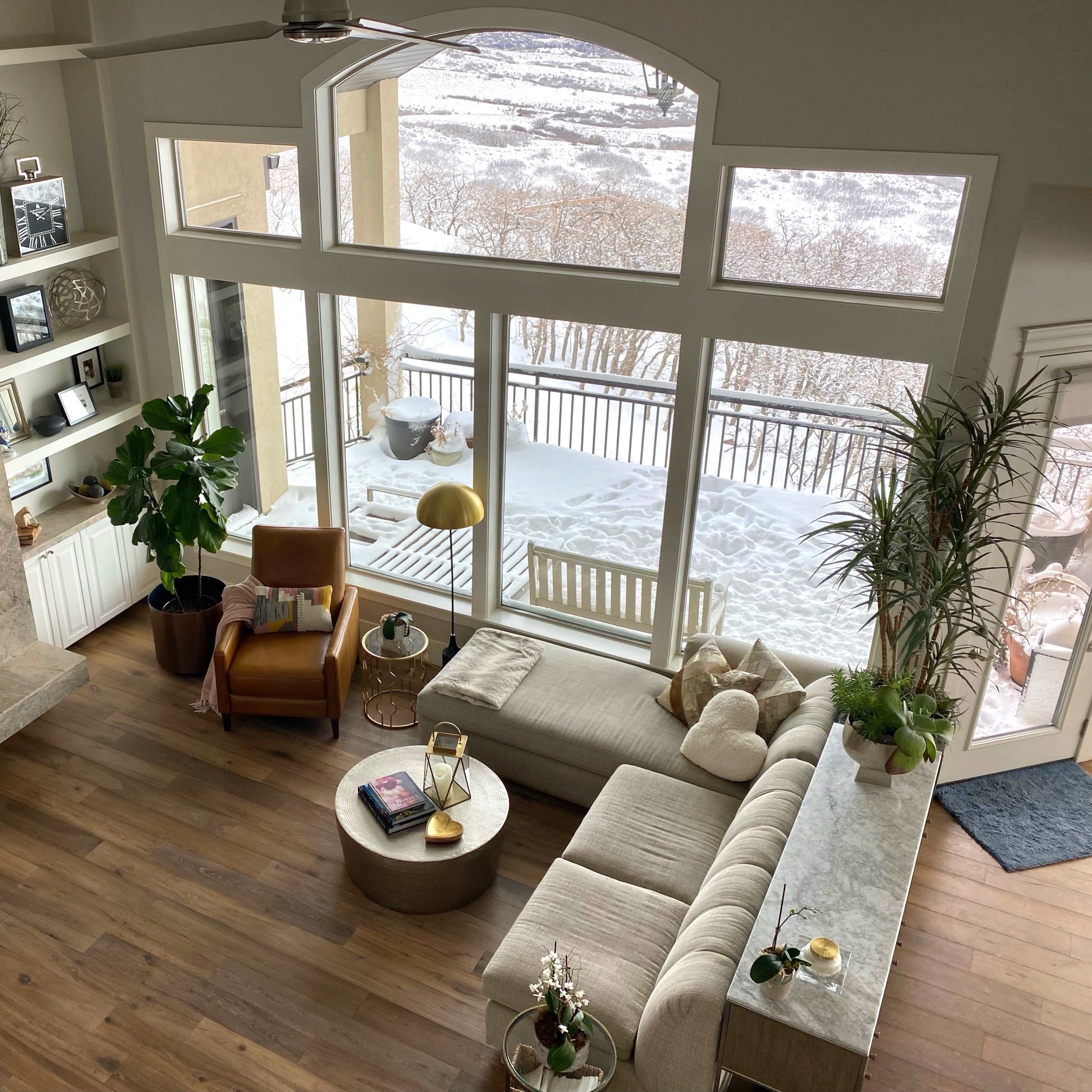 A spacious living room with a large window overlooking a snowy landscape, featuring a modern fireplace, a beige sectional sofa, a wooden accent chair, and decorative plants.