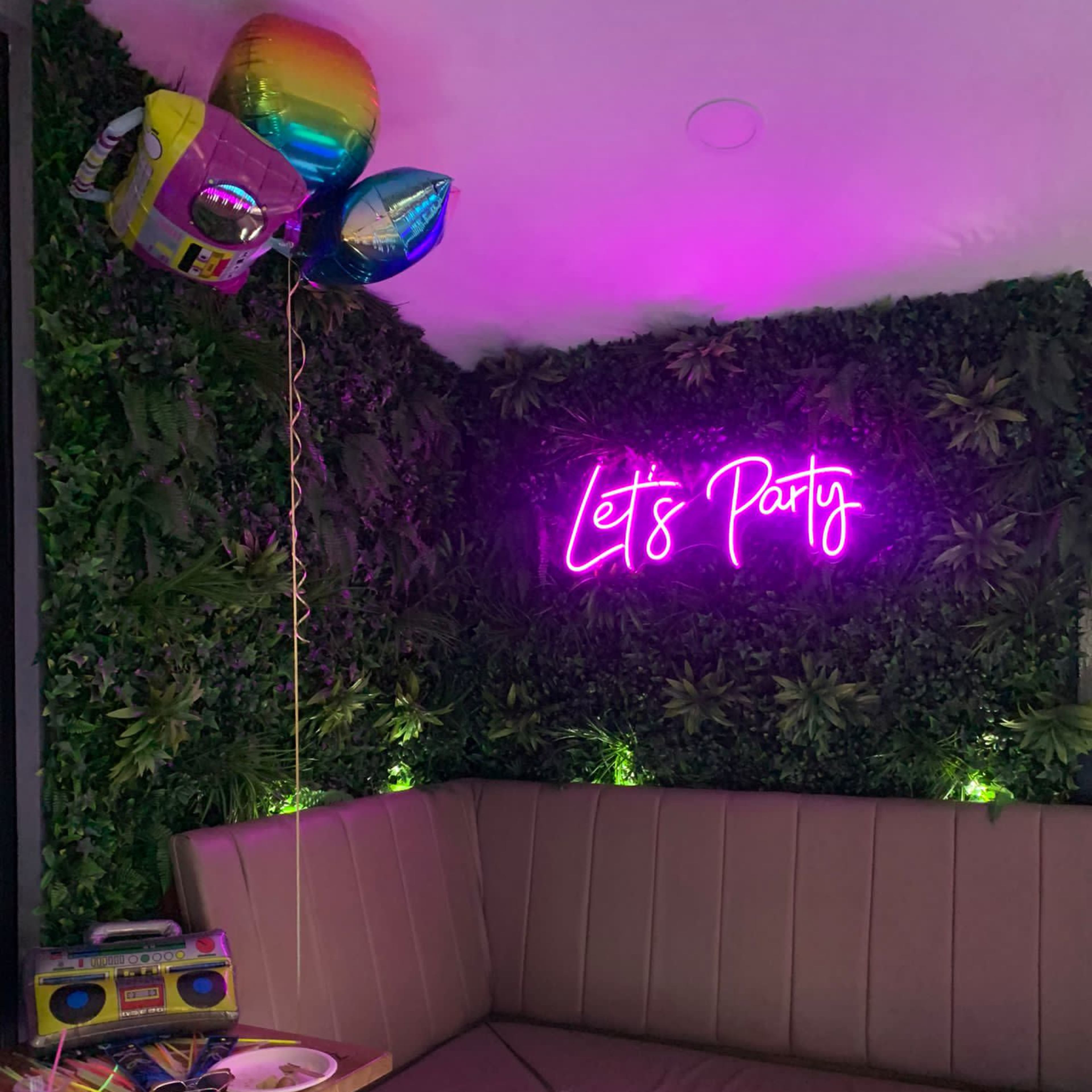 A cozy seating area features a neon sign that reads "Let's Party," surrounded by green plants and colorful balloons.