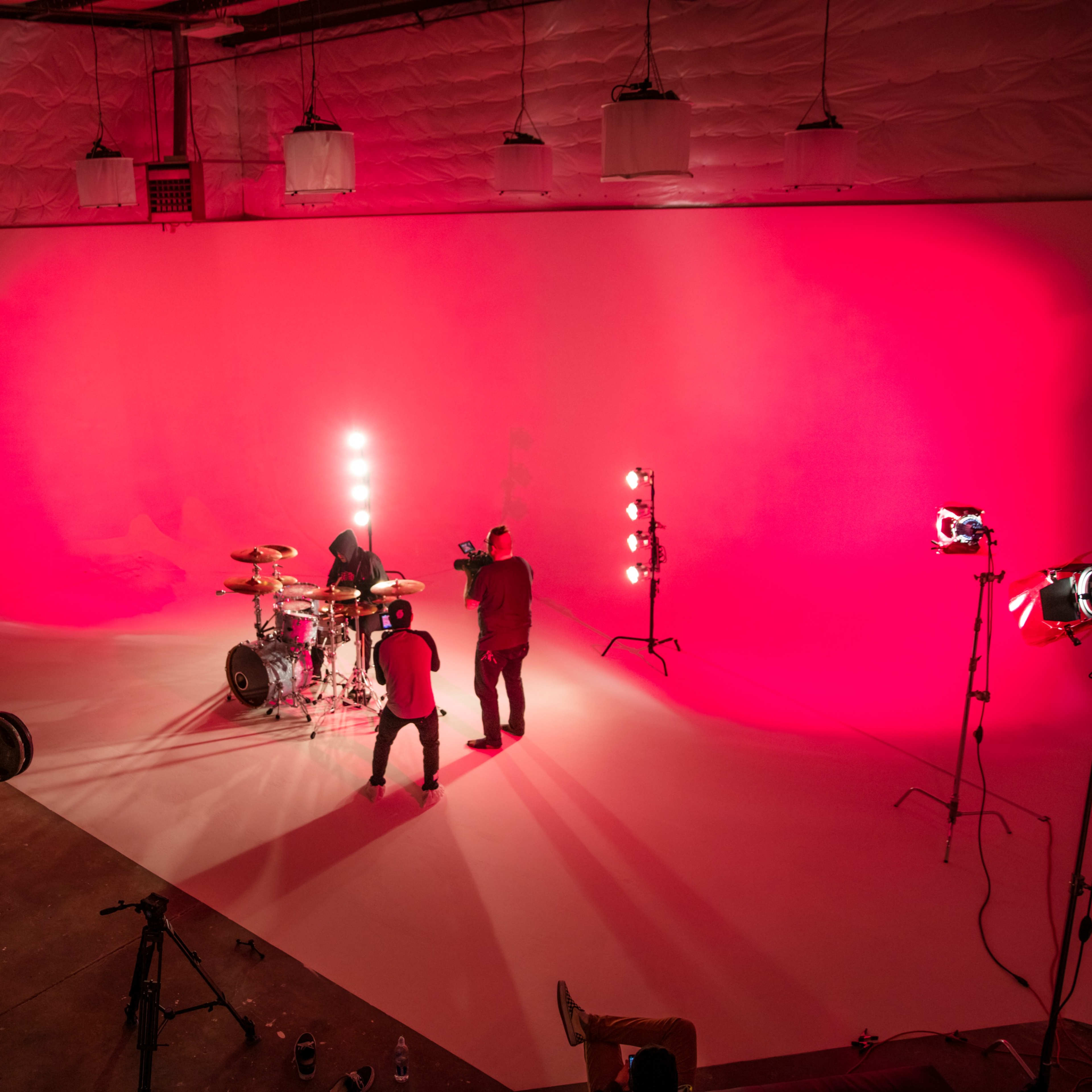 A band performs on a stage with a bright pink backdrop and various lighting equipment set up around them.