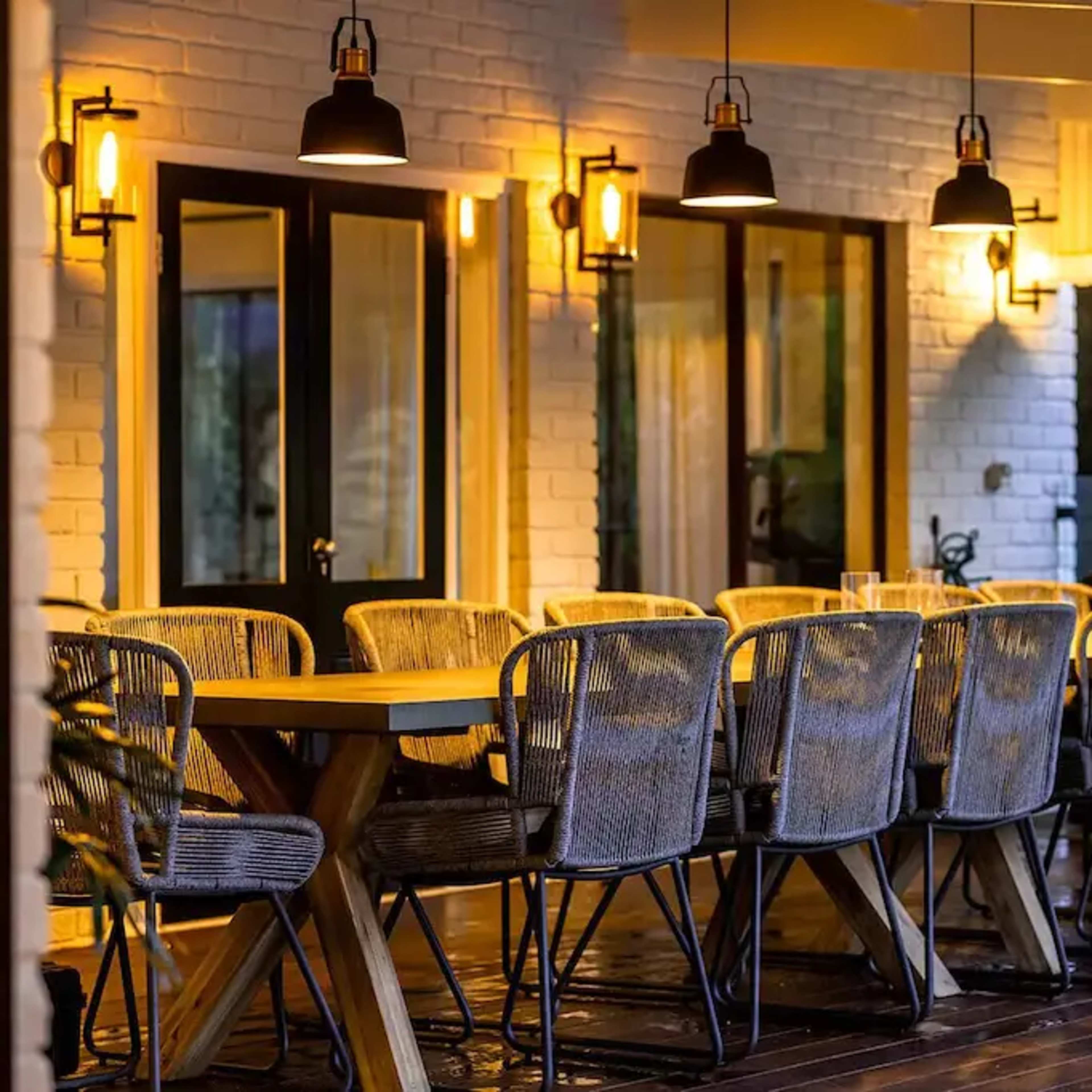 A spacious outdoor dining area features a large wooden table surrounded by wicker chairs, illuminated by pendant lights.
