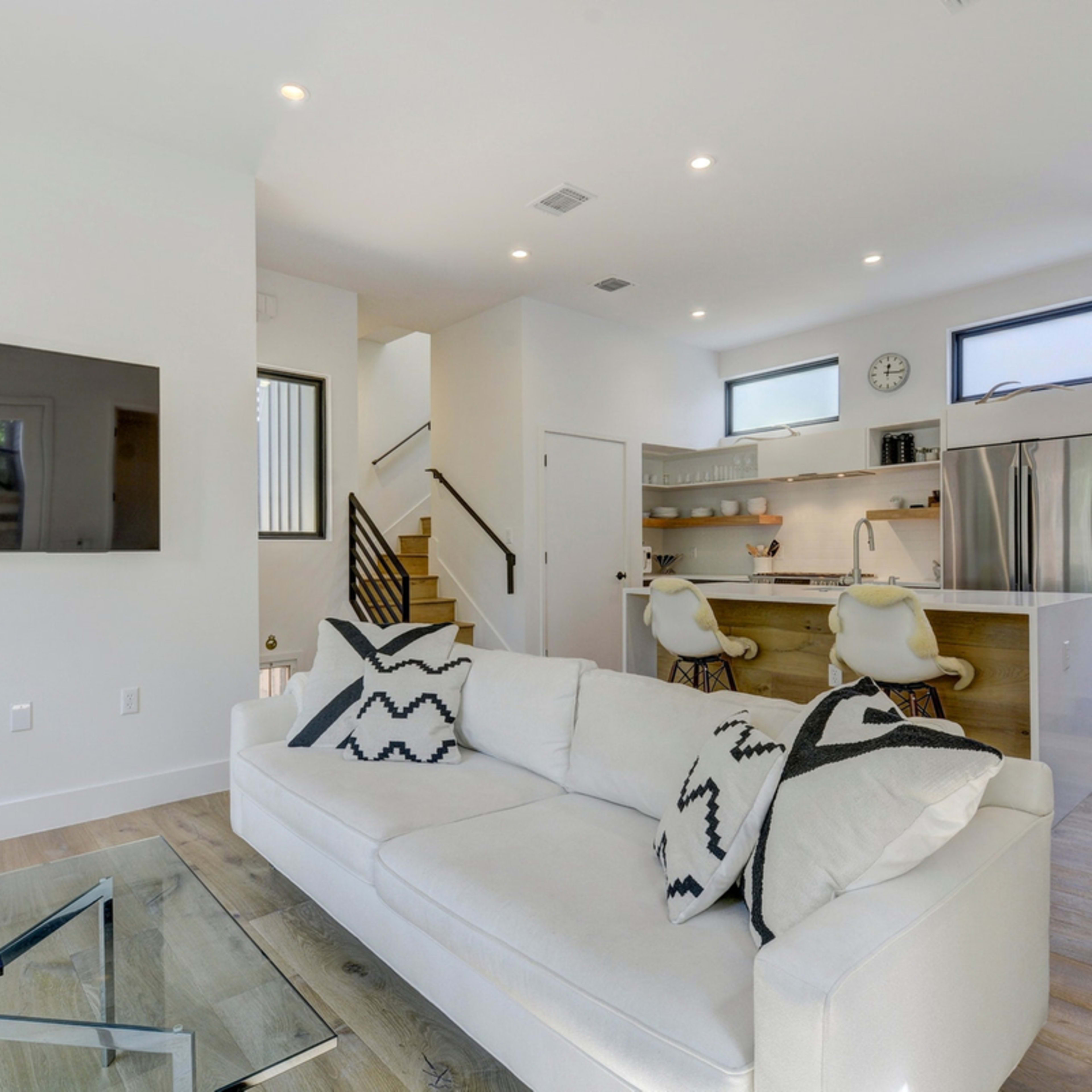 A modern living room features a white sofa with patterned throw pillows, a glass coffee table, and an open kitchen visible in the background.