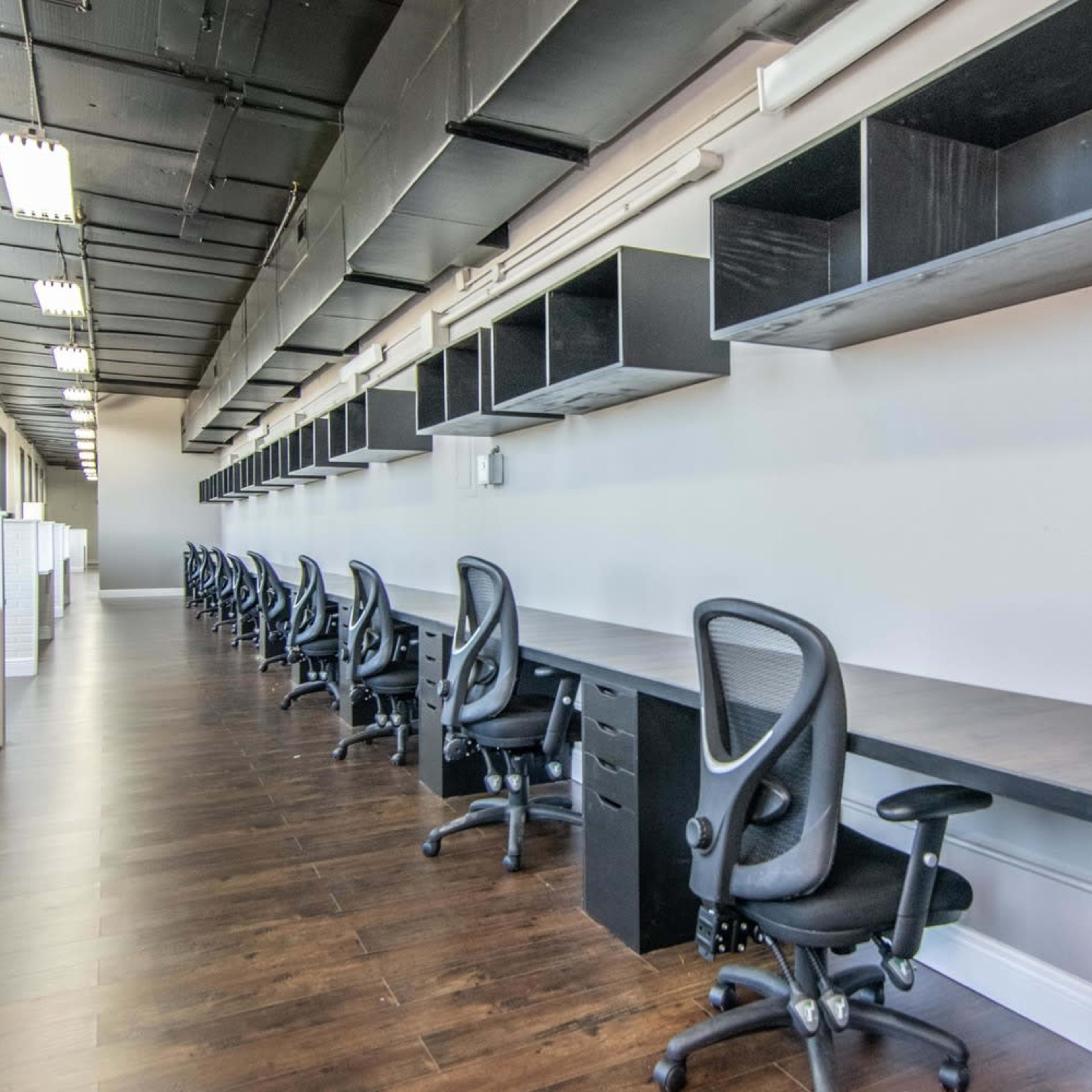 A row of empty office desks with ergonomic chairs is aligned along a spacious, well-lit room featuring overhead shelves.