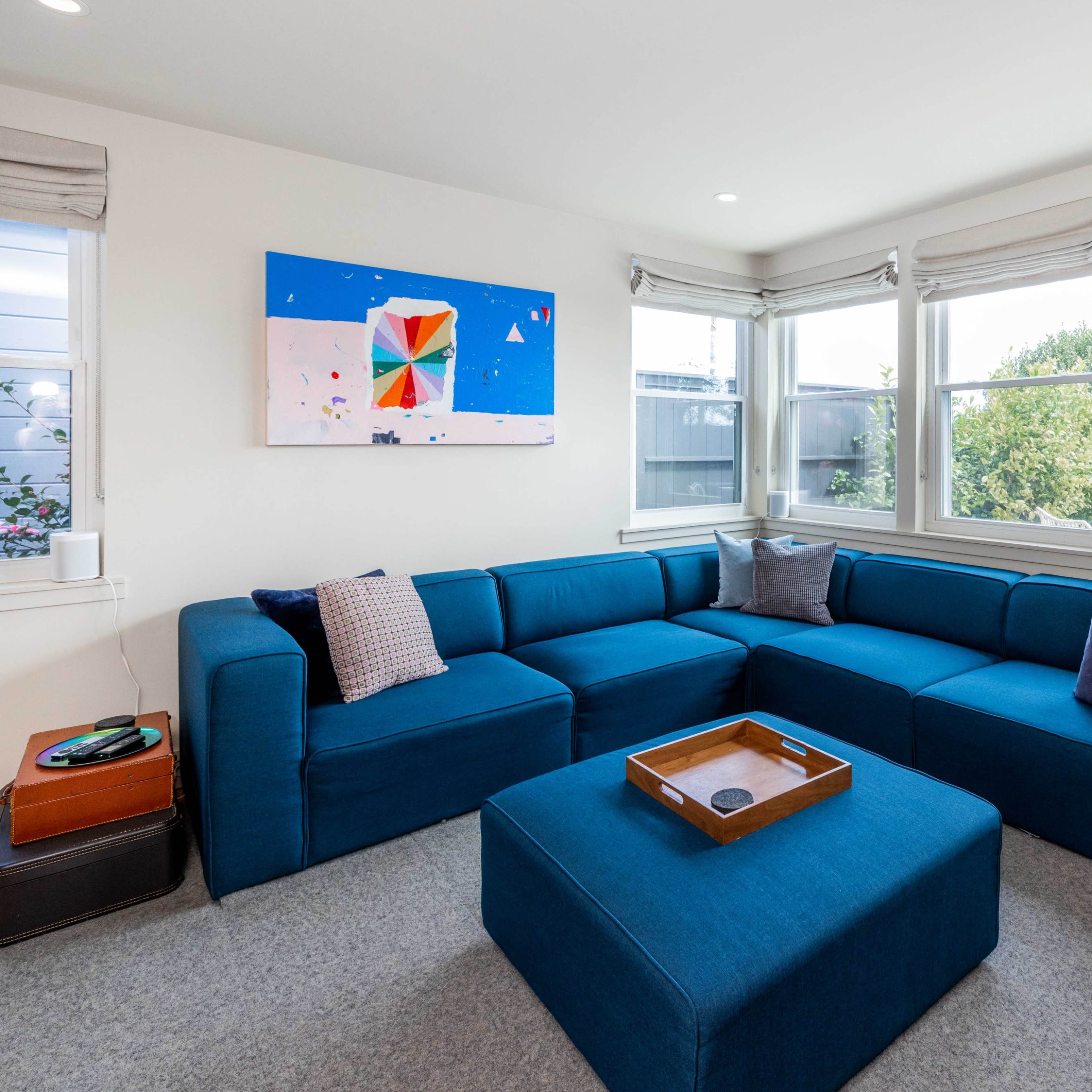 A modern living room with a large blue sectional sofa, an ottoman, and a colorful abstract painting on the wall, illuminated by natural light from several windows.